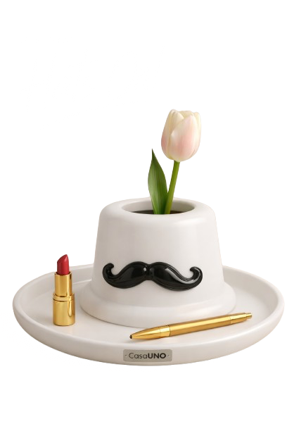 A creative desk setup featuring a white ceramic pot shaped like a hat with a pink tulip and green leaves inside, a black mustache decoration, a gold lipstick, and a gold pen on a round white tray. The background is plain white with a signature or tex
