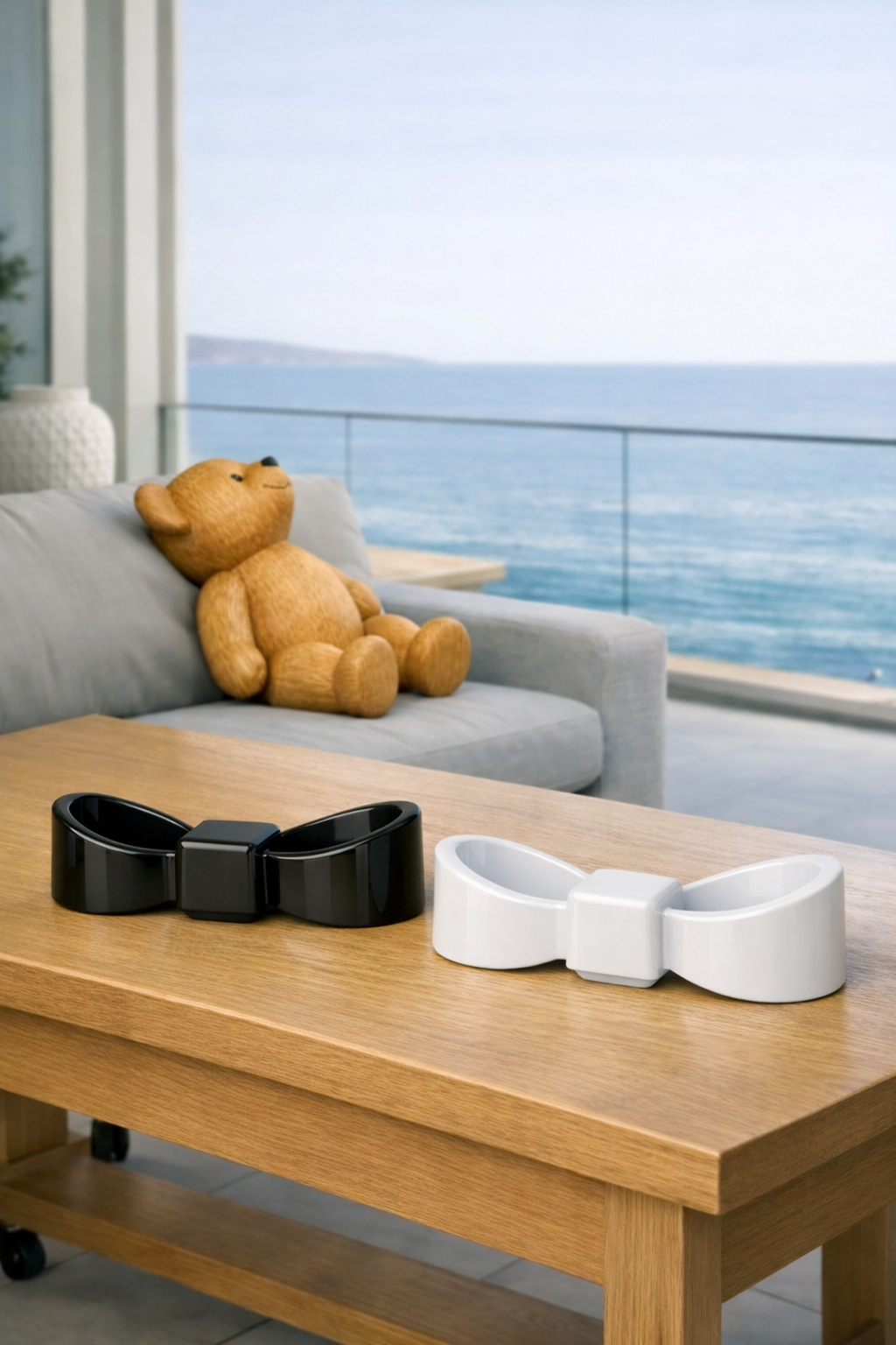 A teddy bear is sitting on a gray sofa overlooking the ocean. On a wooden table in the foreground, there are two bowtie-shaped objects, one black and one white, resembling large hair accessories or decorative pieces.