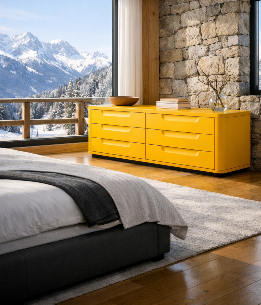 Modern bedroom with a large window showing a snowy mountain landscape, a yellow dresser with a wooden bowl, books, and a glass vase with branches, a bed with white and black bedding, and a beige rug on wooden floor.