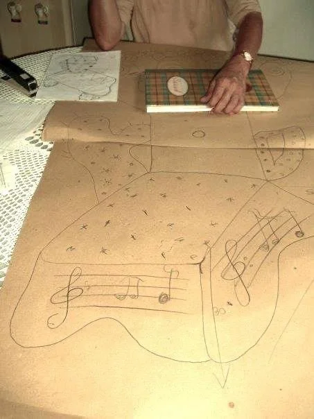 A person is standing behind a wooden table with handmade musical notes and symbols drawn on a large sheet of paper. The person has their hand on a smaller, checkered notebook and is wearing a beige shirt and a watch.