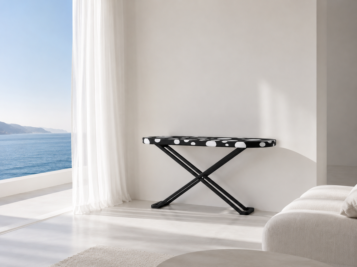 A black and white polka dot ironing board in a minimalist white room near a large window with sheer curtains, overlooking a view of the ocean and mountains.