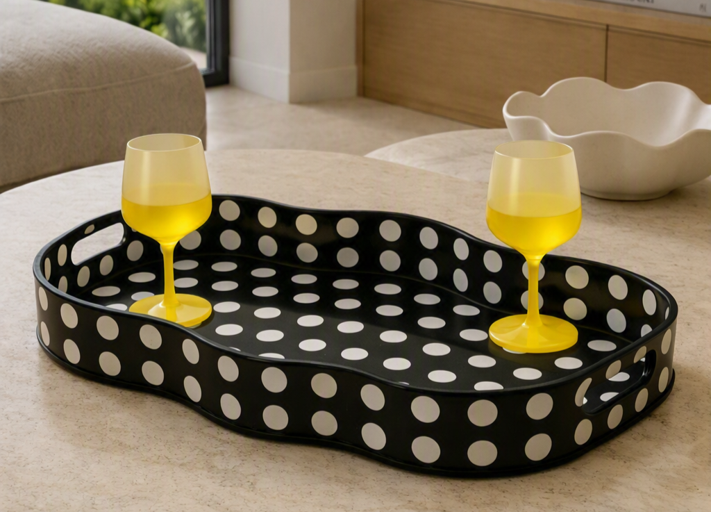 Black tray with white polka dots holding two yellow wine glasses filled with yellow drink, on a white table in a room with beige furniture and a decorative white bowl.