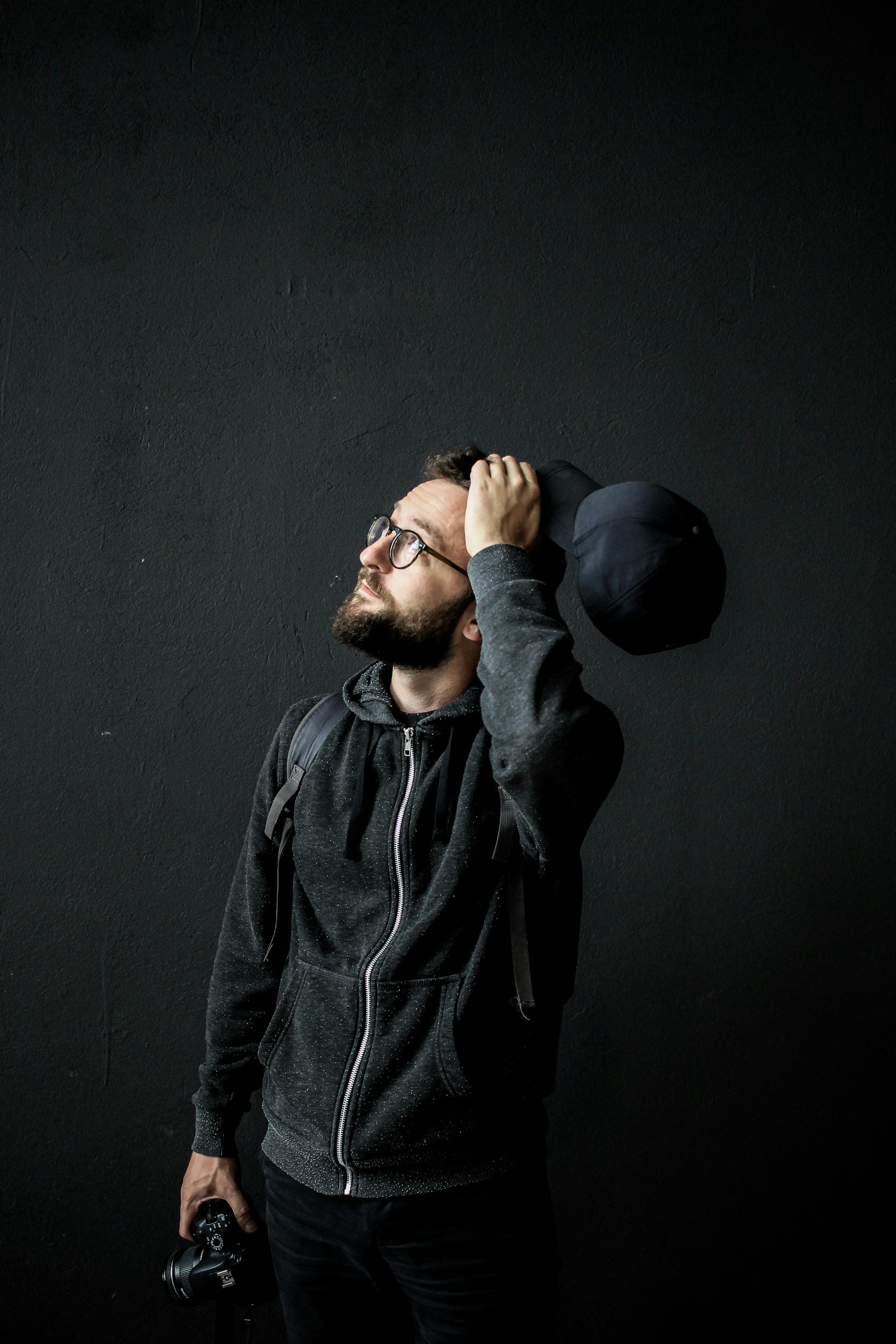A man with glasses and a beard stands against a dark wall, holding a black cap on his head with his right hand and holding a camera in his left hand.