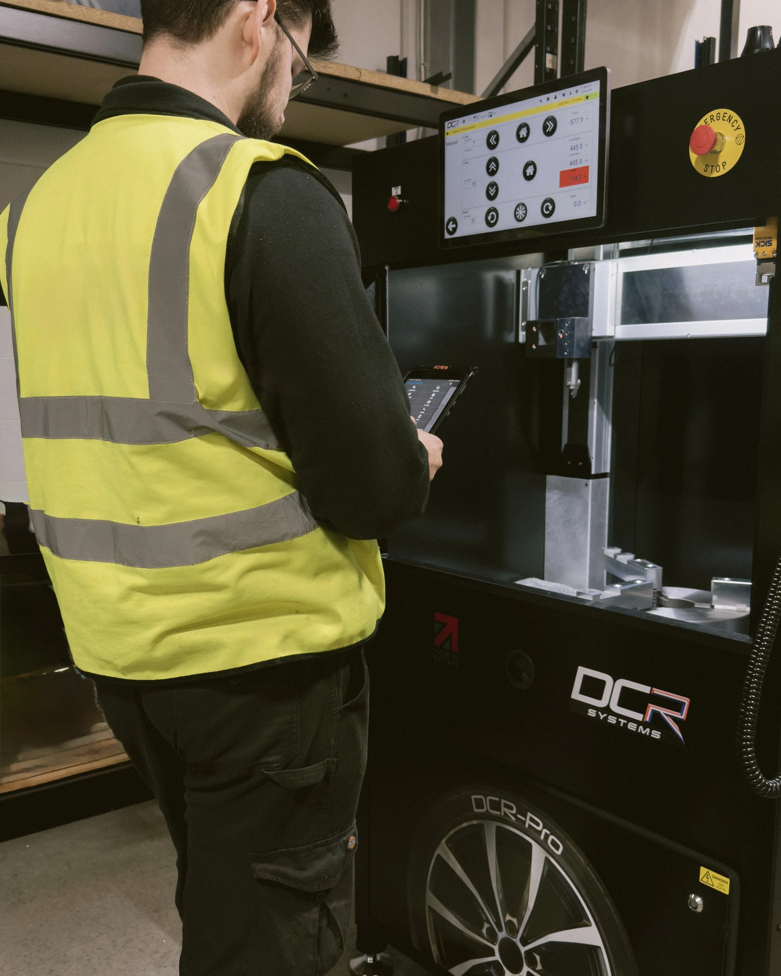 A person wearing a yellow safety vest and black clothing is operating a DCR systems machine in a workshop or industrial setting. The person is holding a tablet and looking at the machine's digital screen, which displays various data and controls.