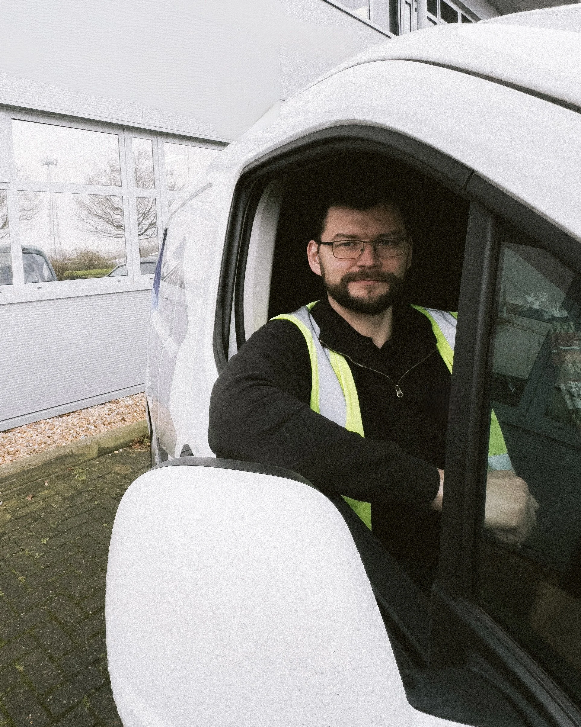A man with glasses and a beard sitting in the cockpit of a small, modern vehicle, wearing a black shirt and a neon yellow safety vest, outside a building with large windows.