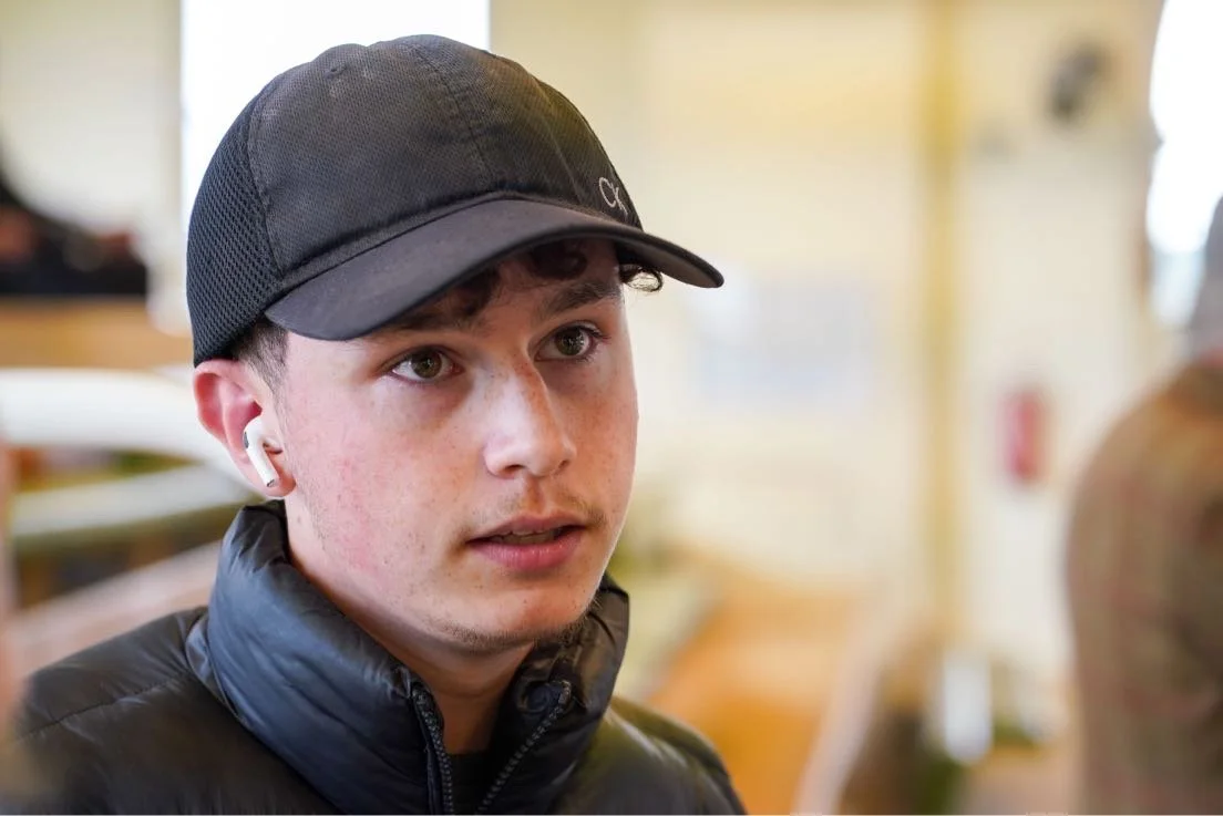 Close-up of young man wearing black cap, black jacket, and white wireless earbuds, indoors with blurred background.