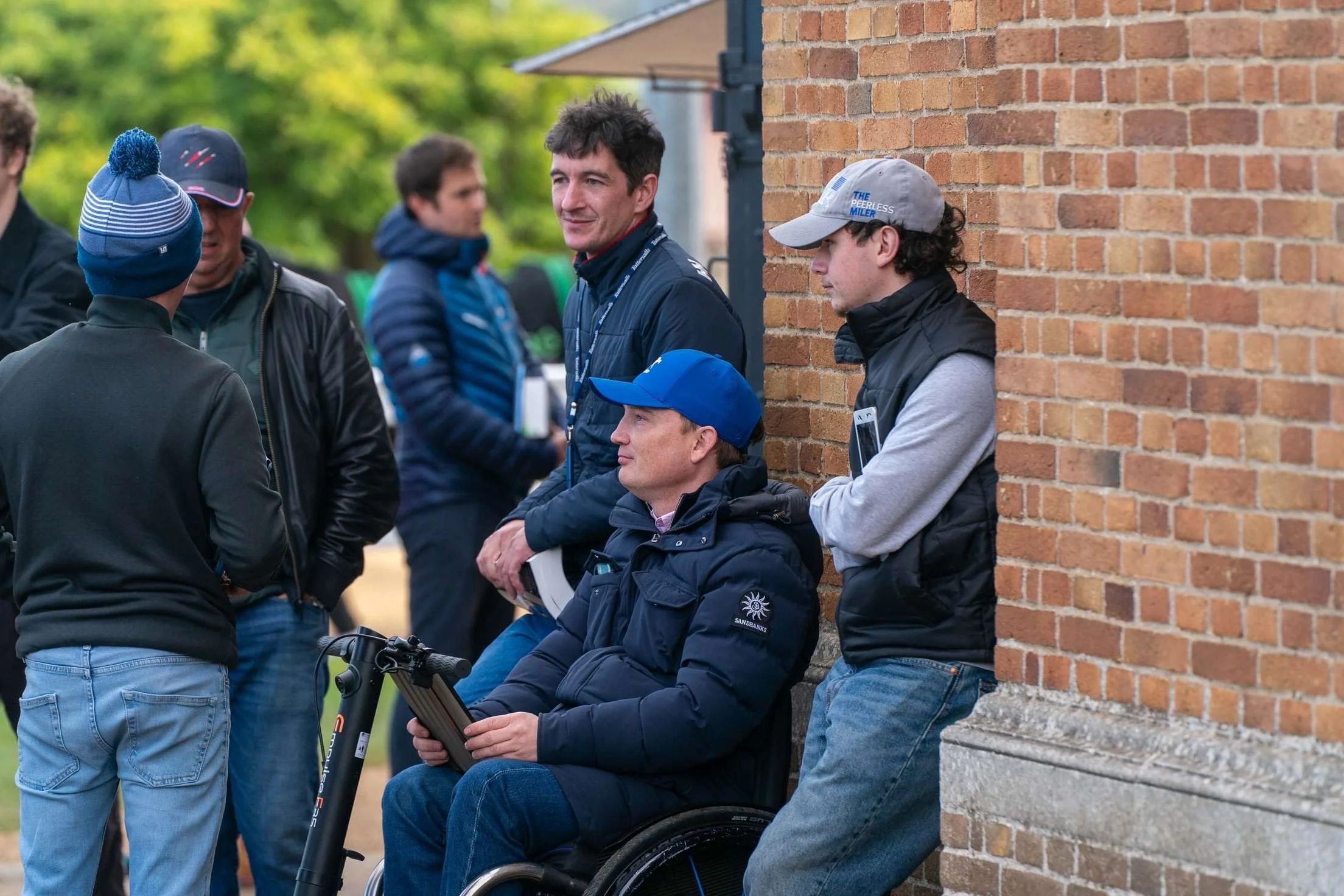 Group of men conversing outdoors, one in a wheelchair holding a tablet, with others standing around, near a brick wall.