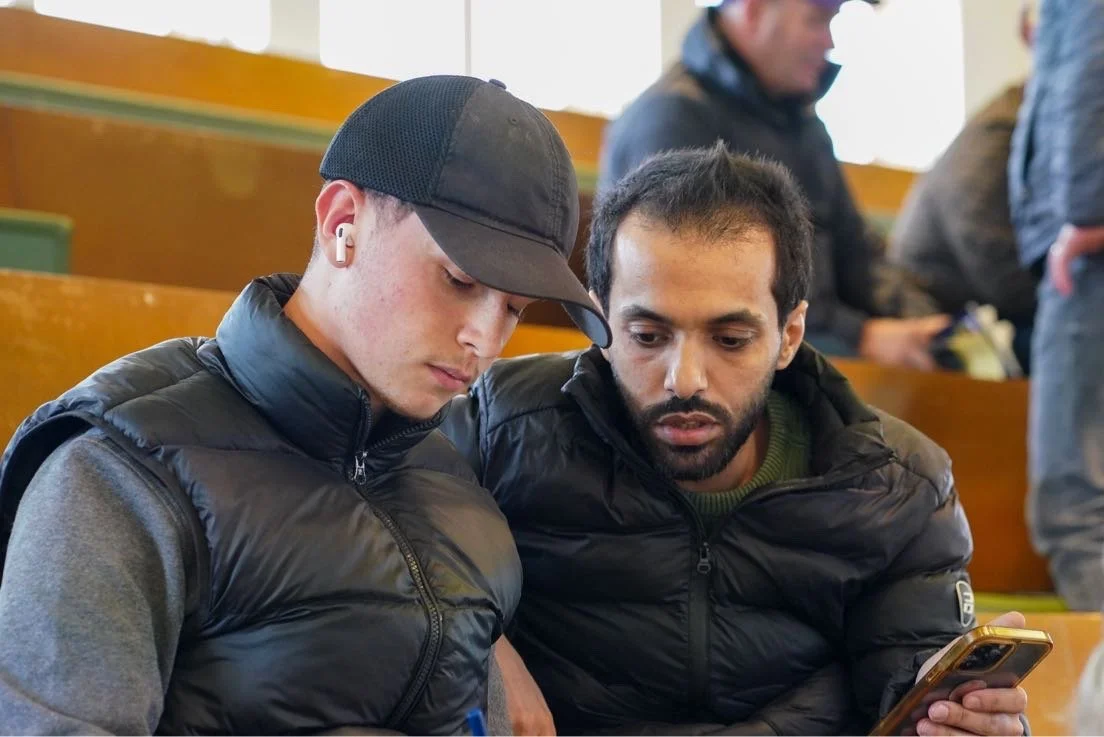 Two young men sitting closely in a classroom or auditorium, looking at a smartphone together, with other people seated in the background.