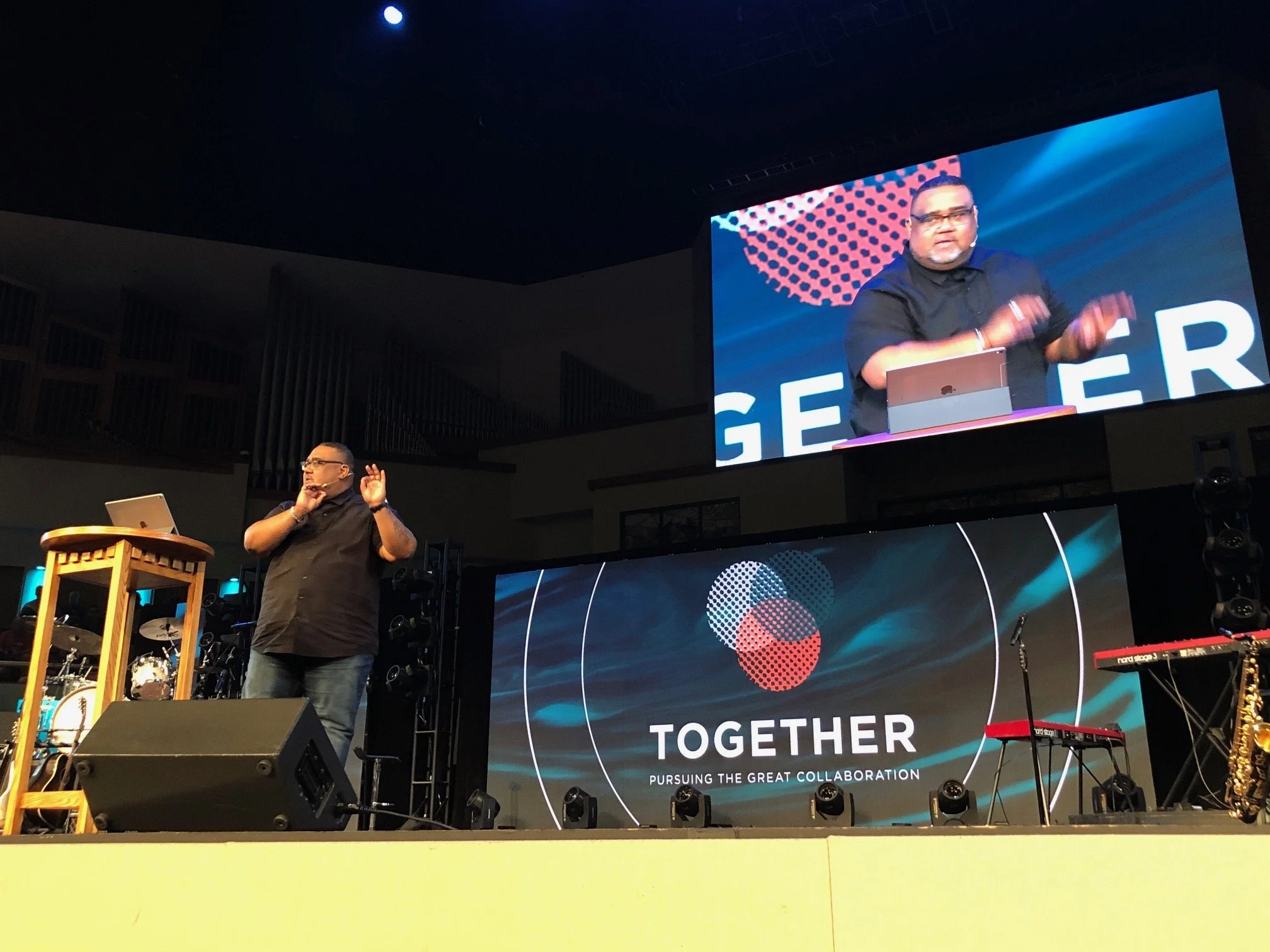 A man speaking on stage at a conference titled 'TOGETHER' with a subtitle 'Pursuing the Great Collaboration.' A large screen behind him displays his image, and the stage has musical instruments including a keyboard, microphone, and drums.