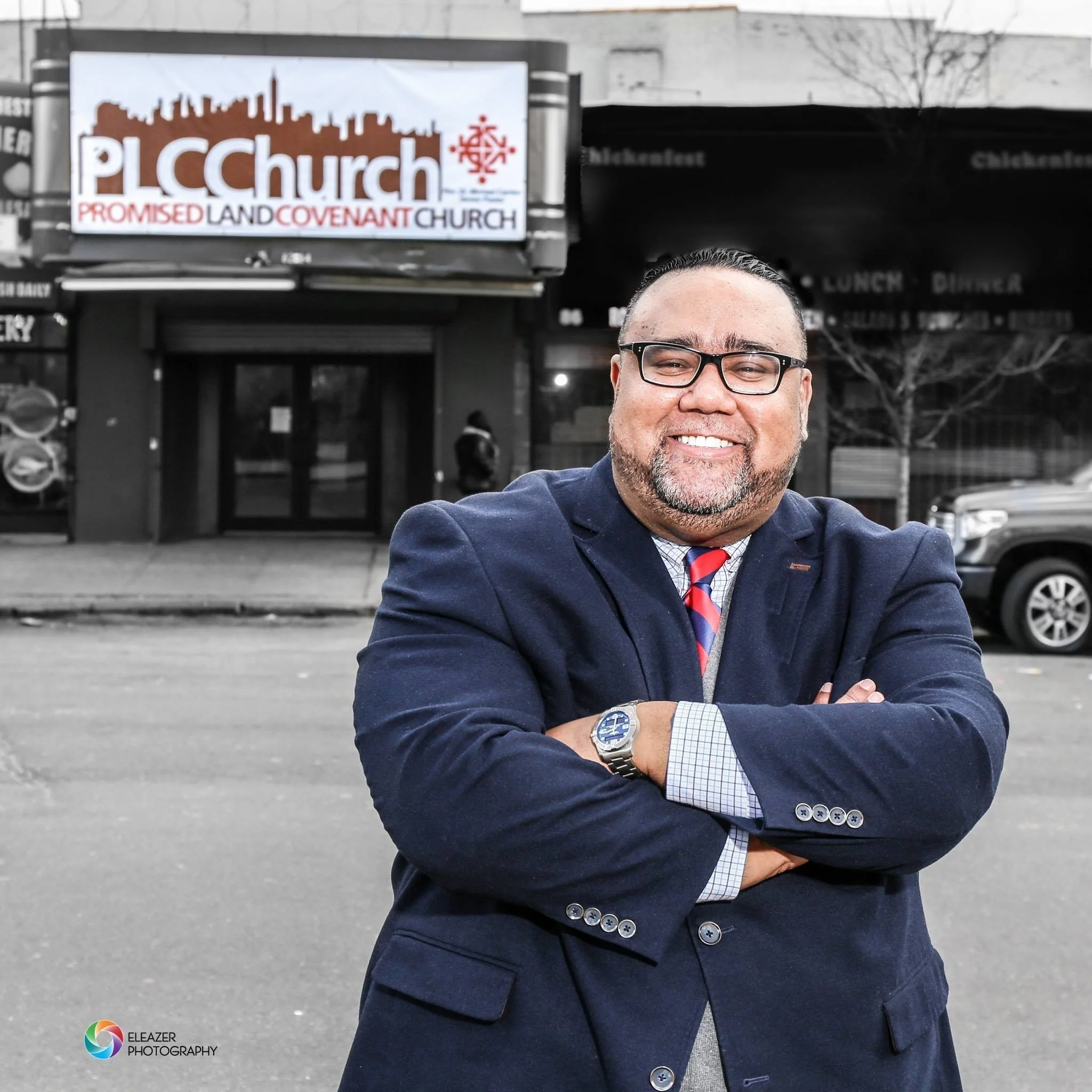 A man in a navy blazer with crossed arms, wearing glasses, smiling in front of a black and white background that features a sign for PLCC Church, Promised Land Covenant Church, with a person walking in the background and parked cars to the side.