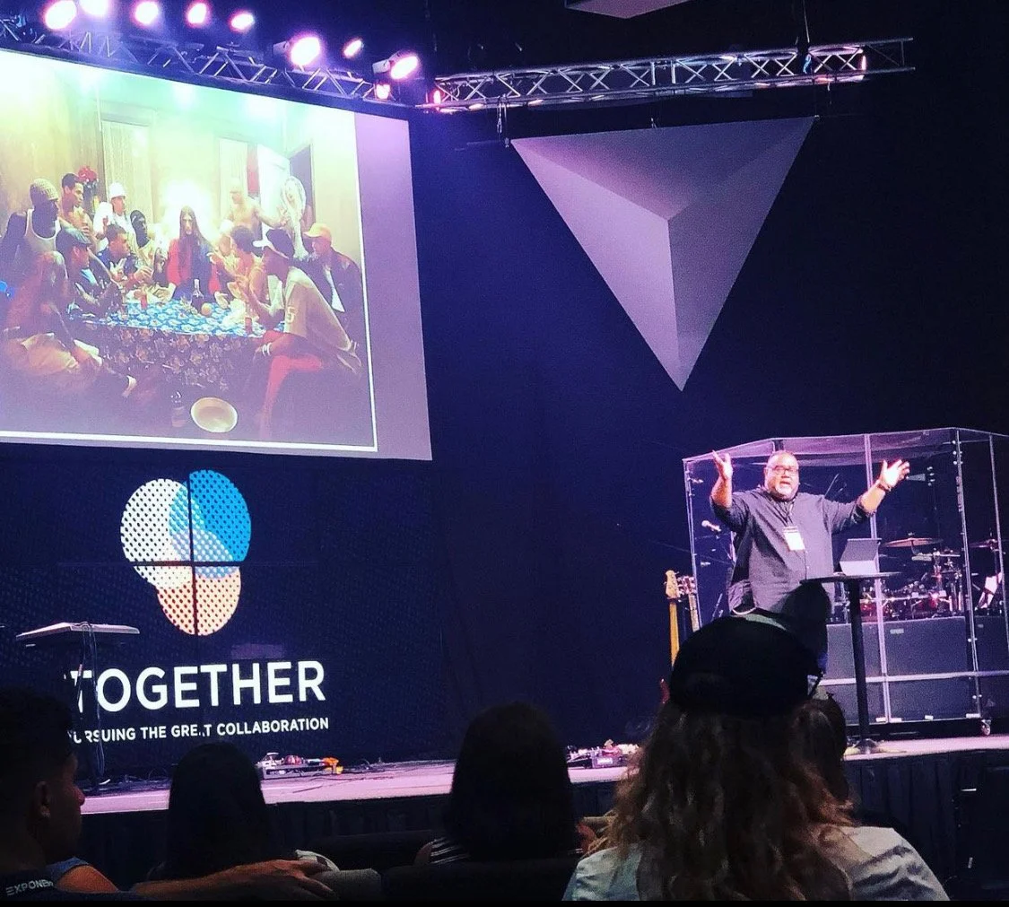 A speaker stands on stage behind a transparent lectern with a group of people seated in front. A large screen displays a photo of a group of people engaging in a collaborative activity. The stage features lights and geometric decorations, with the word "TOGETHER" and the tagline "using the great collaboration" projected on the screen.