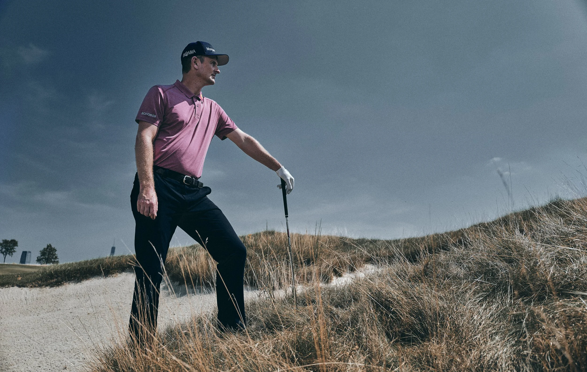 A man wearing a purple polo shirt, black pants, a golf cap, and white gloves playing golf outdoors on a cloudy day, standing on a golf course with grassy dunes.