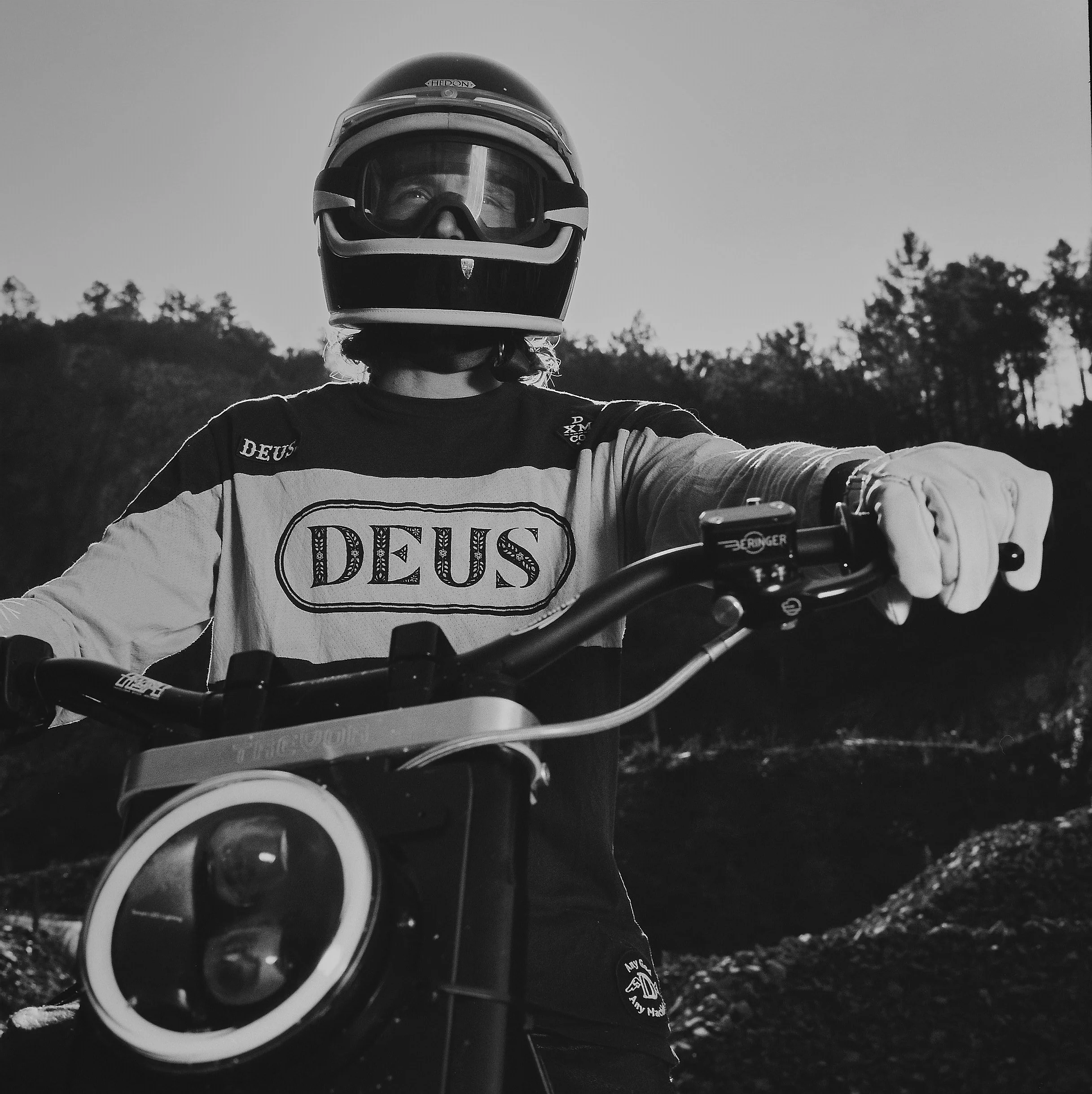 A person wearing a motorcycle helmet with goggles, riding a motorcycle outdoors at dusk or dawn, with trees in the background.