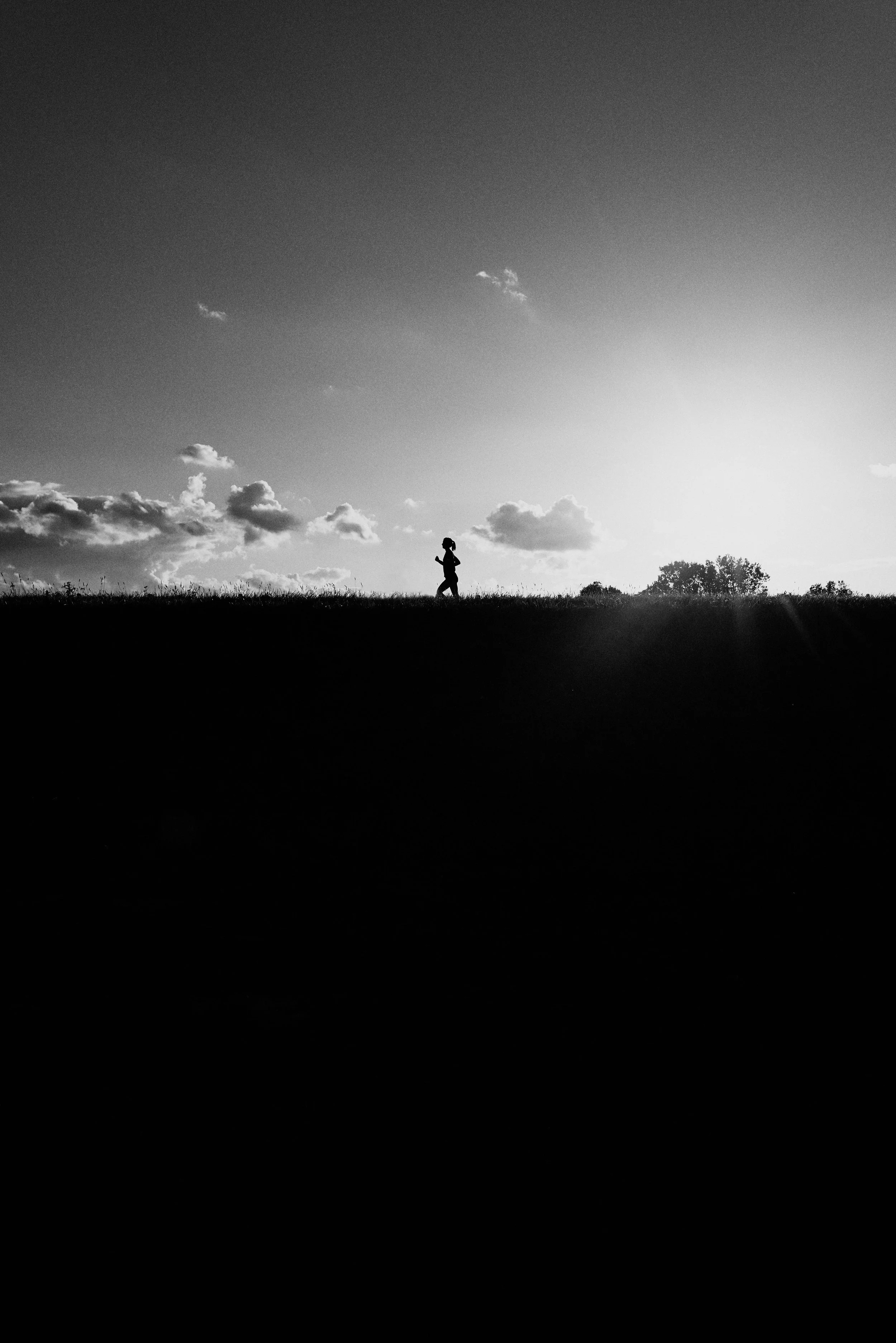 Silhouette of a person jogging on a grassy hill at sunset, with clouds and sky in the background, black and white image.