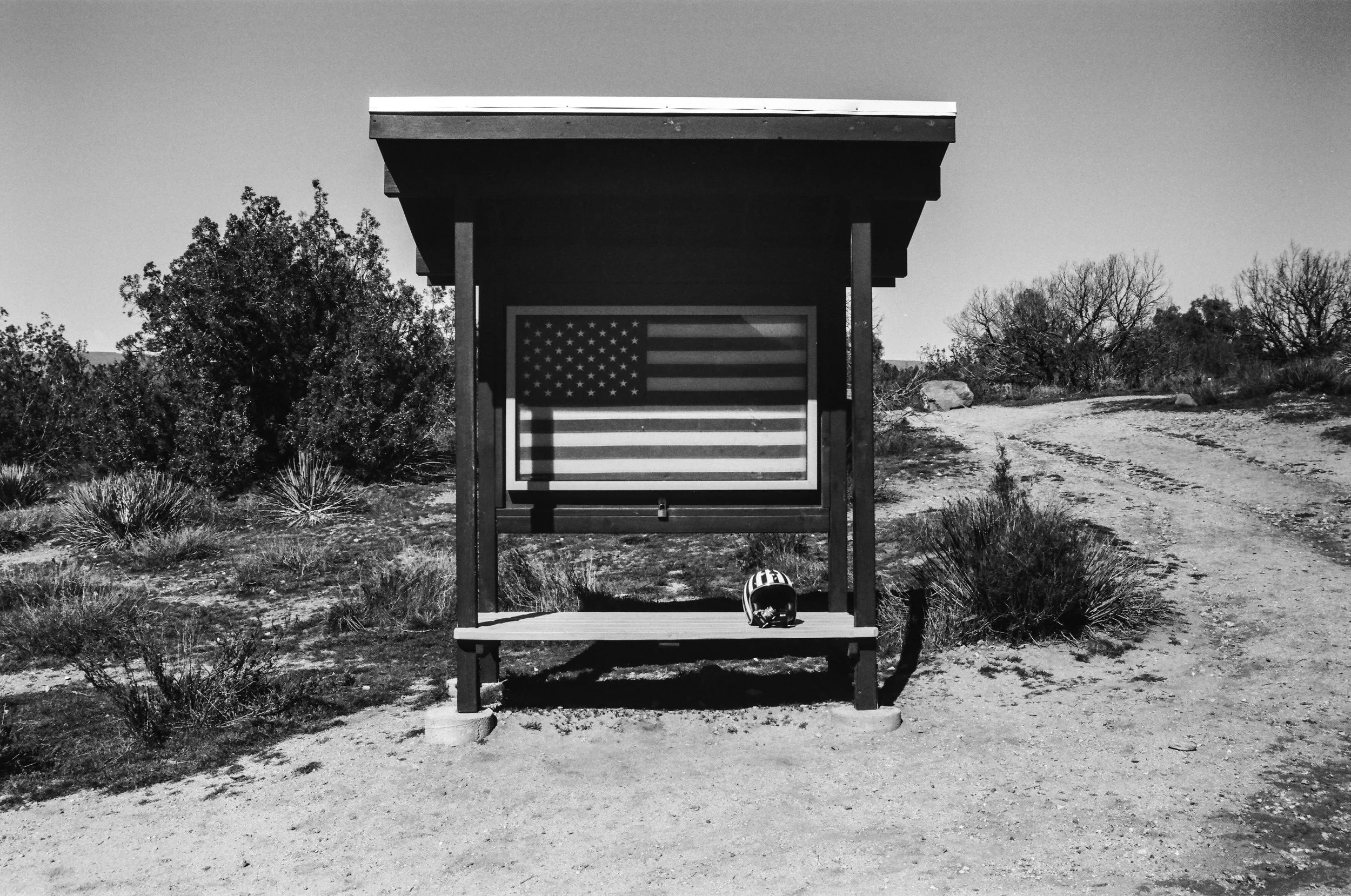 A black and white photo of a small wooden shelter with an American flag on the back wall. There is a helmet placed on the bench underneath the shelter. The shelter is situated in a desert landscape with bushes, rocks, and a dirt road. Trees and open 