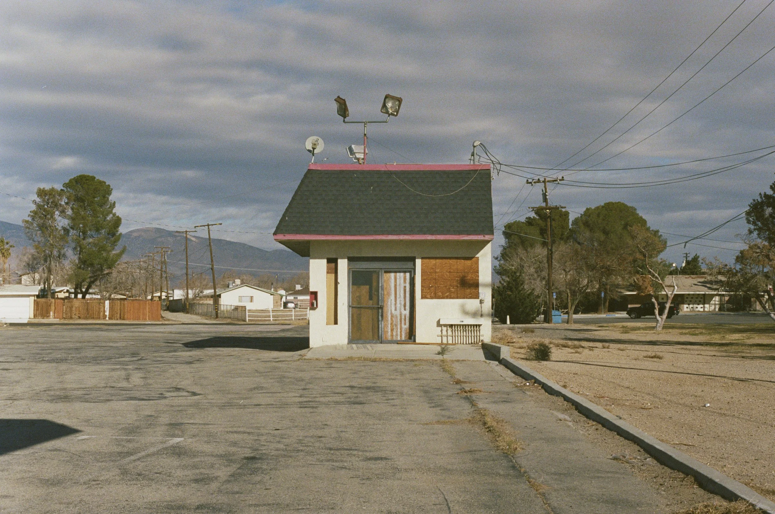 A small, empty building with boarded windows and a door, located in a parking lot. The building has a dark shingled roof with pink trim and mounted outdoor lights, satellite dish, and antennas on top. Surrounding the lot are trees, utility poles, and a backdrop of mountains with a cloudy sky.
