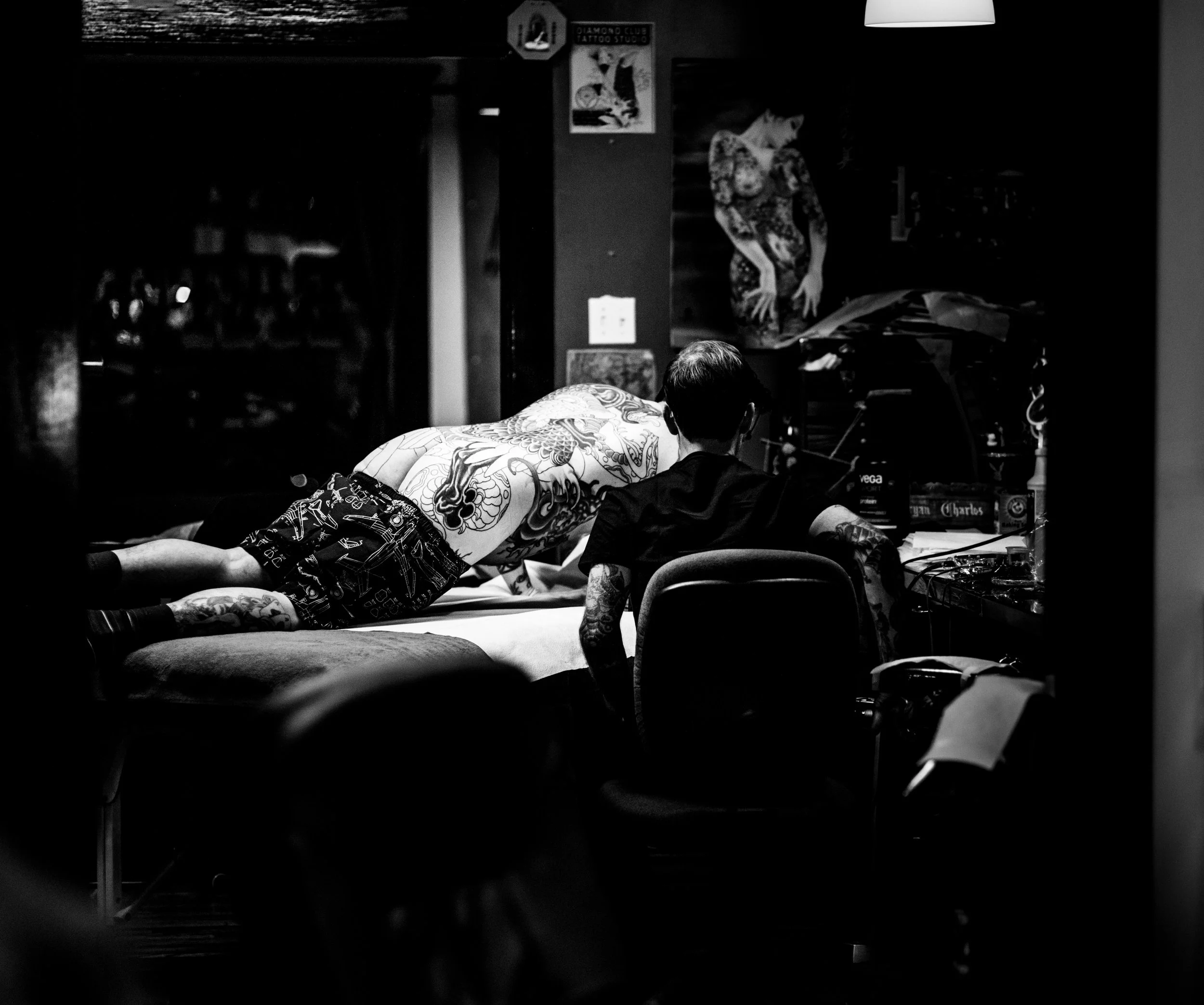 A person with tattoos lying face down on a bed, wearing black shorts with a pattern, in a dimly lit room with posters on the wall that include a cat and a sign for a tattoo studio.