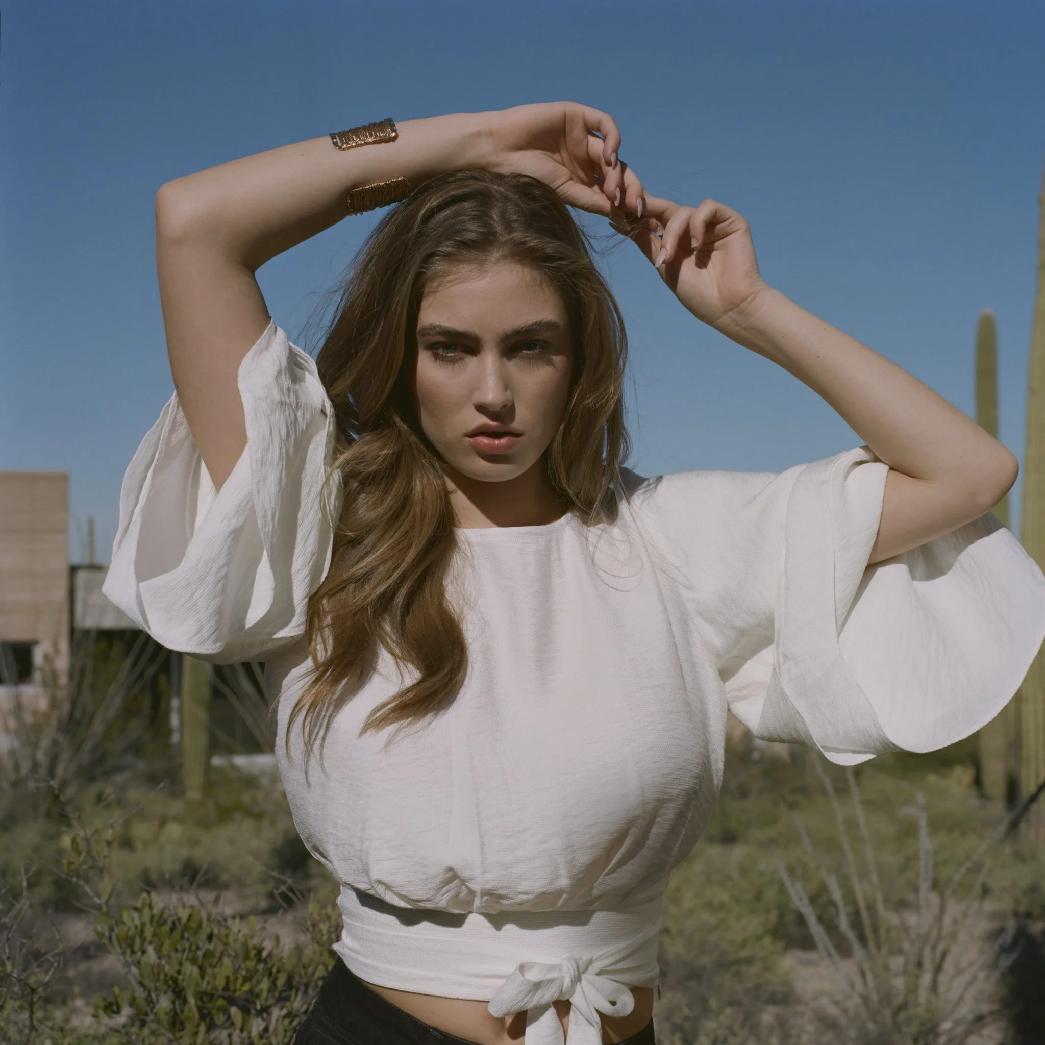 A woman with long wavy hair wearing a white tied-up shirt with rolled-up sleeves, standing outdoors against a clear blue sky, with a desert plant in the background.