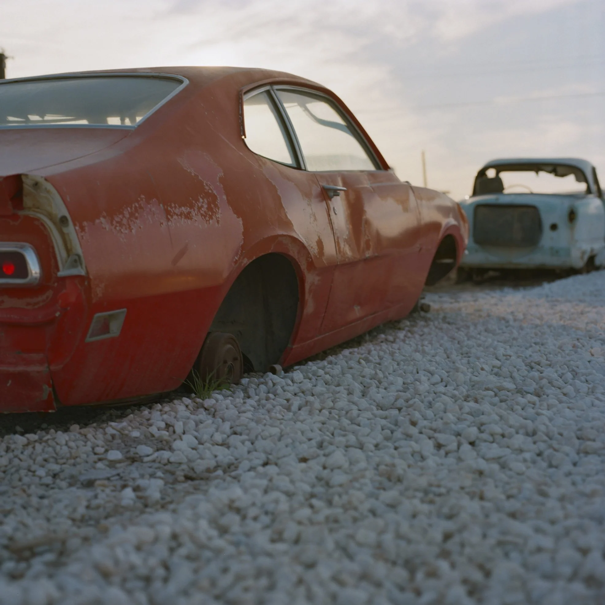 A red vintage car with peeling paint and missing tail light, parked on gravel alongside other old cars during sunset.