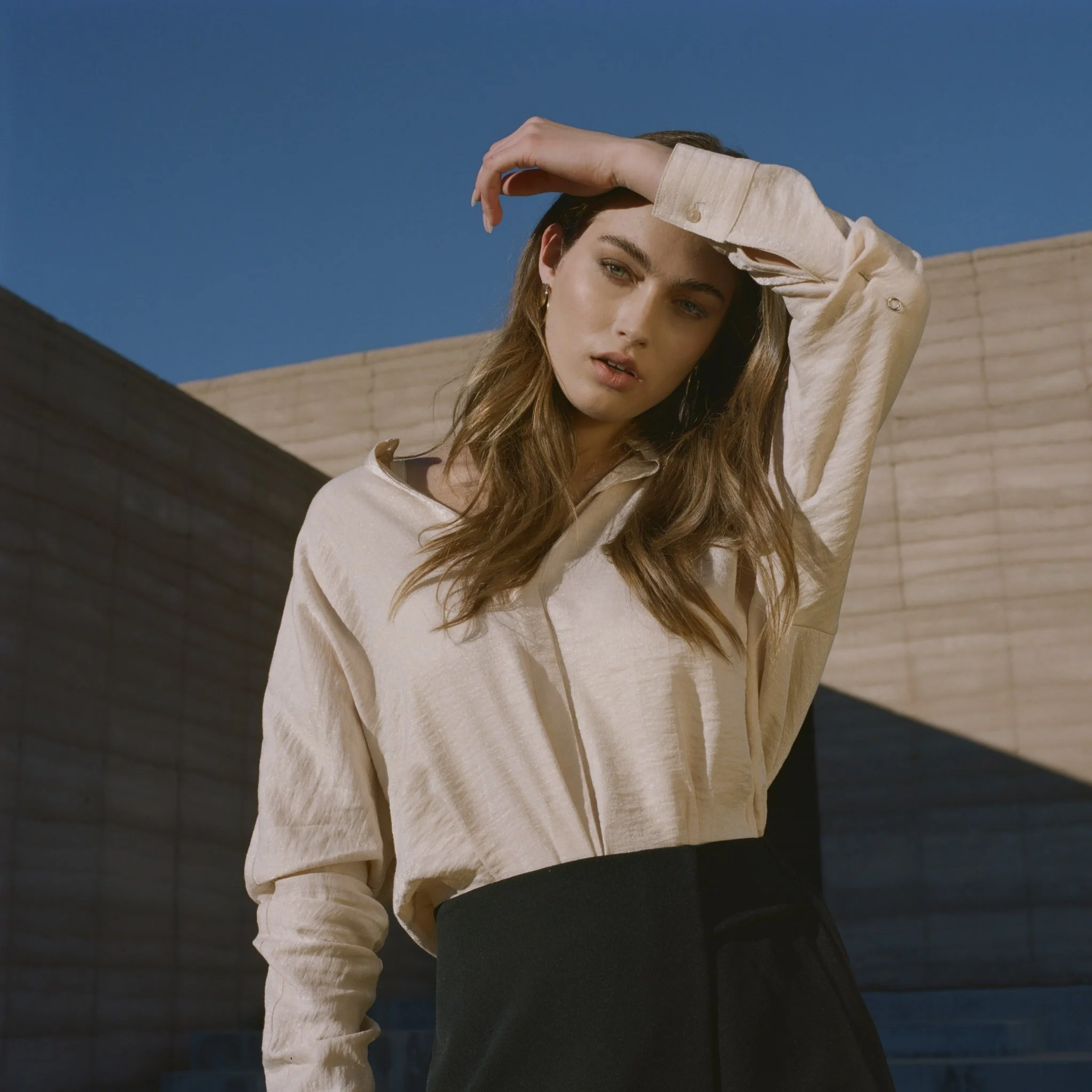 A young woman with fair skin and long wavy brown hair, wearing a beige satin blouse with button details on the cuffs and a black skirt, standing outdoors against a blue sky and modern wooden structures.