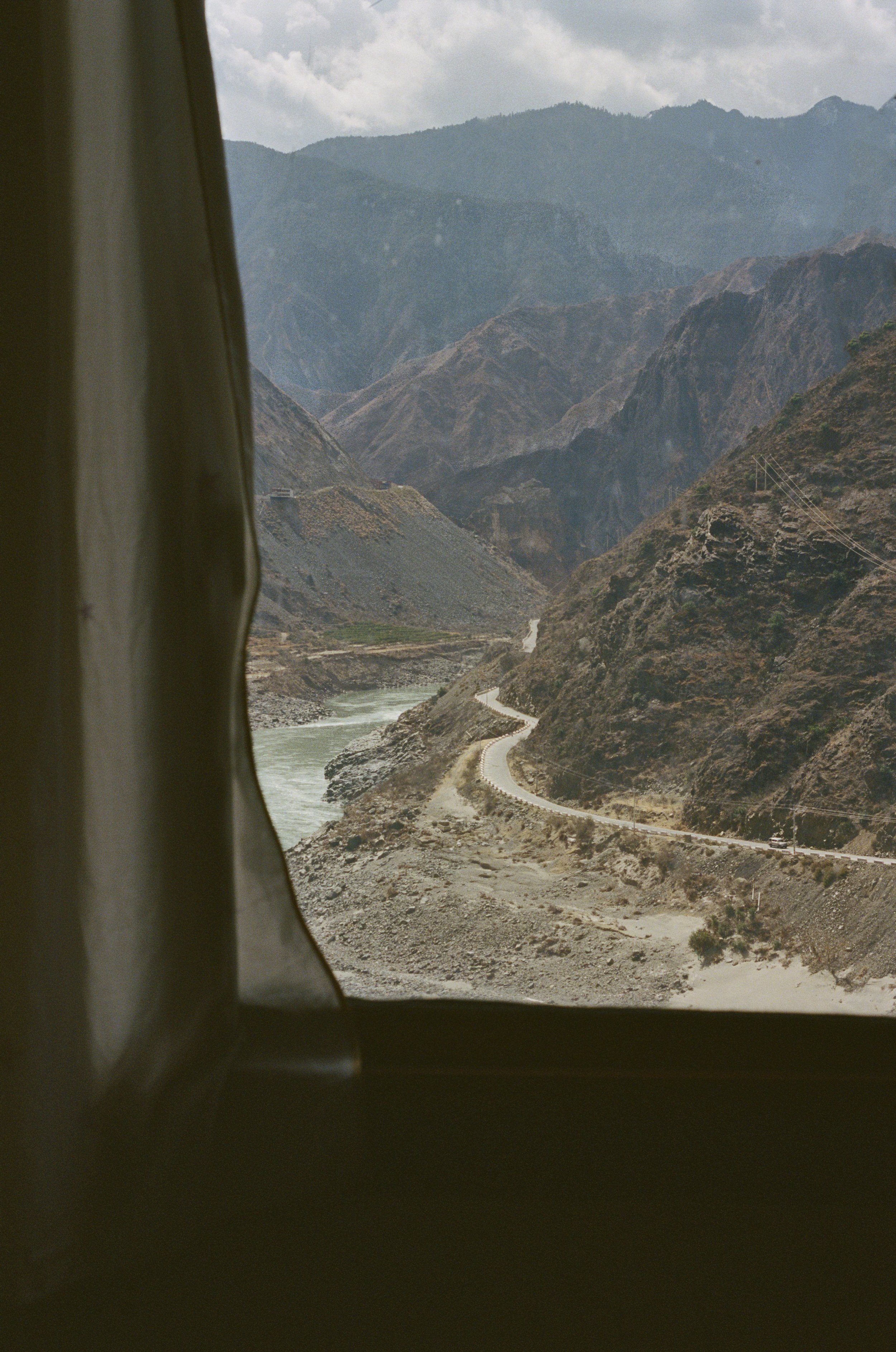 View of winding mountain road next to river, with rugged hills and mountains in the background, seen through a vehicle window with curtain on the side.