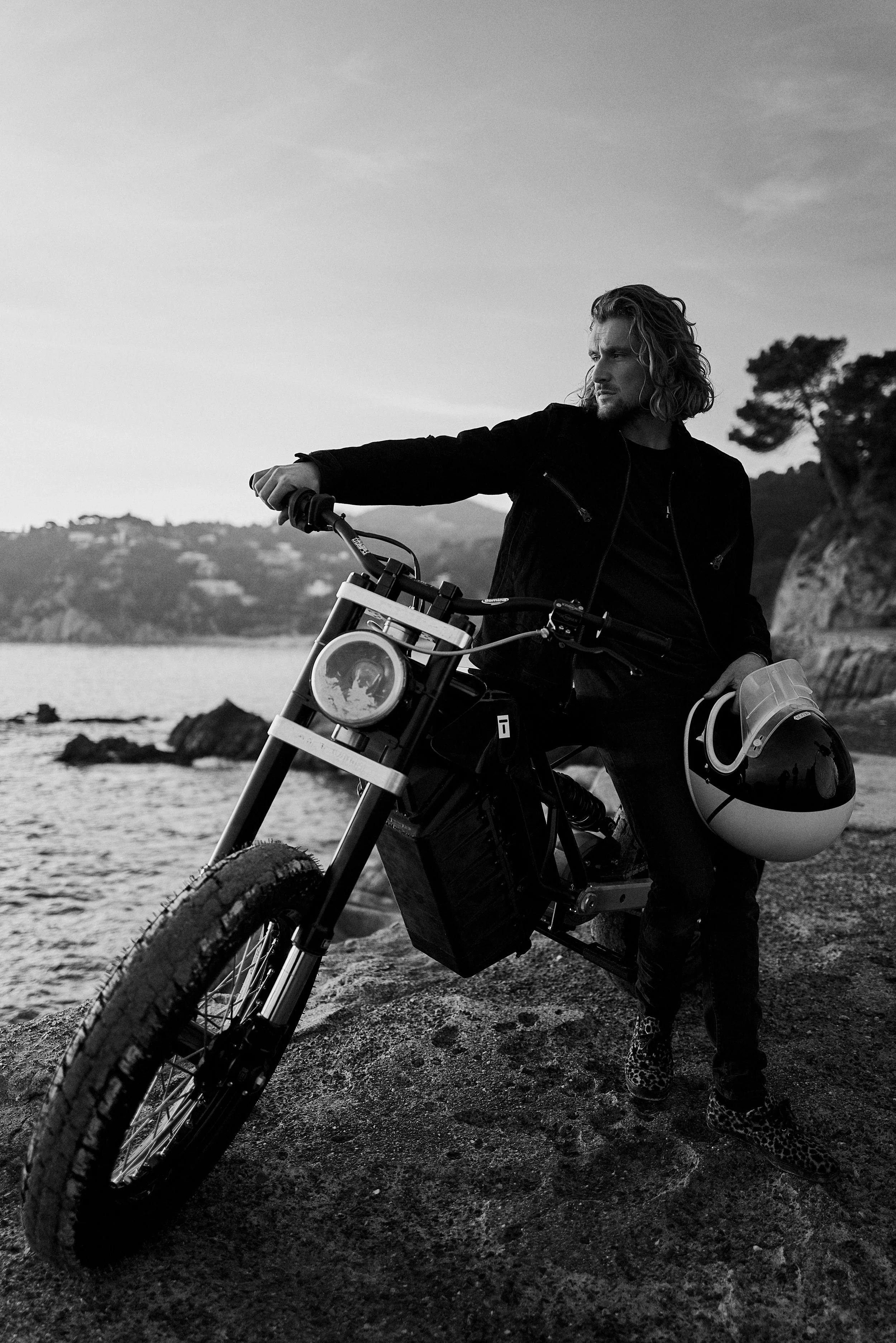 A man with long hair wearing a dark jacket stands beside a motorcycle on a rocky shore, holding a helmet. The background features water, distant hills, and a tree, captured in black and white.