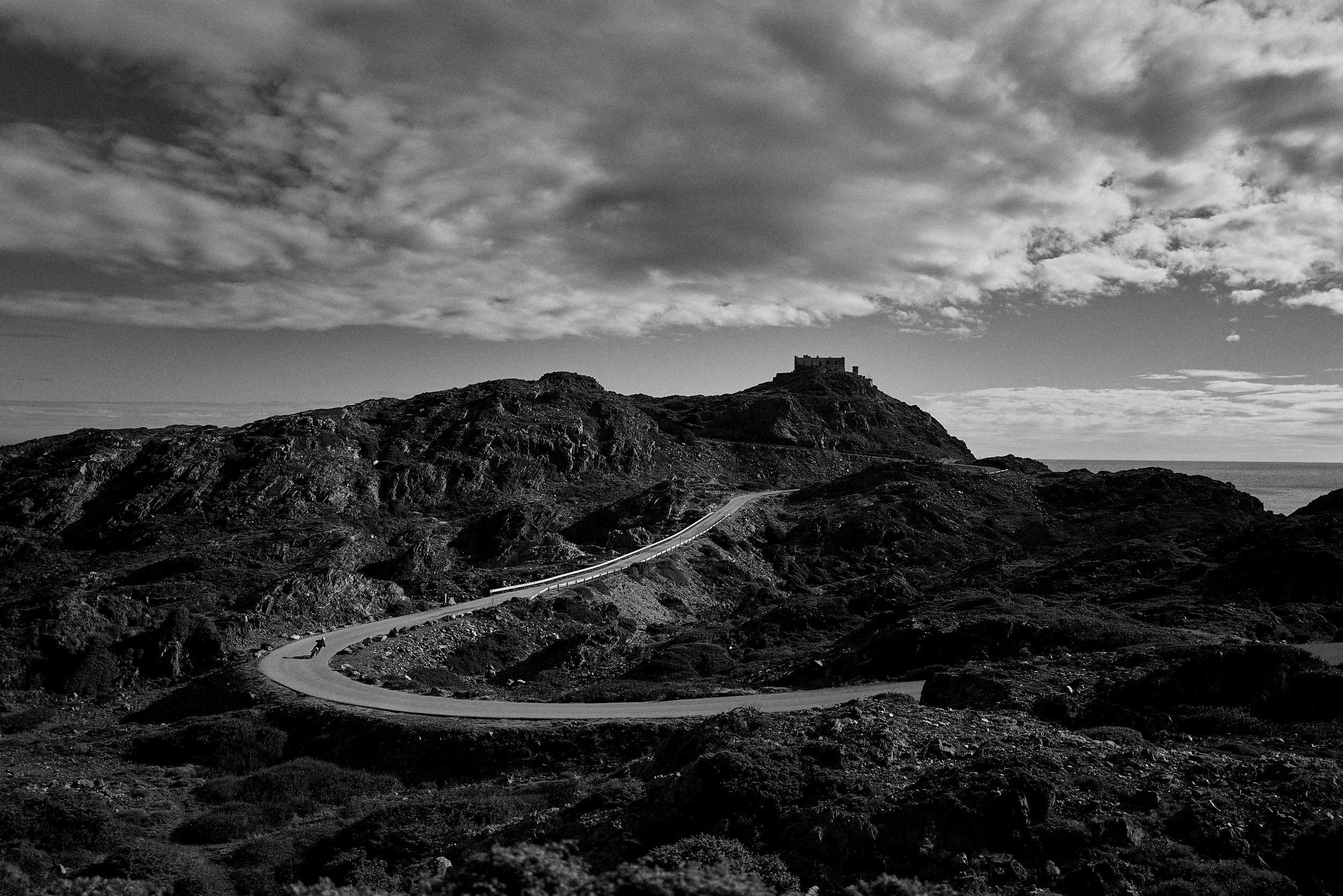 A black and white photograph of a coastal landscape with rugged hills and a winding road leading to a castle or fortress on top of a hill under a cloudy sky.