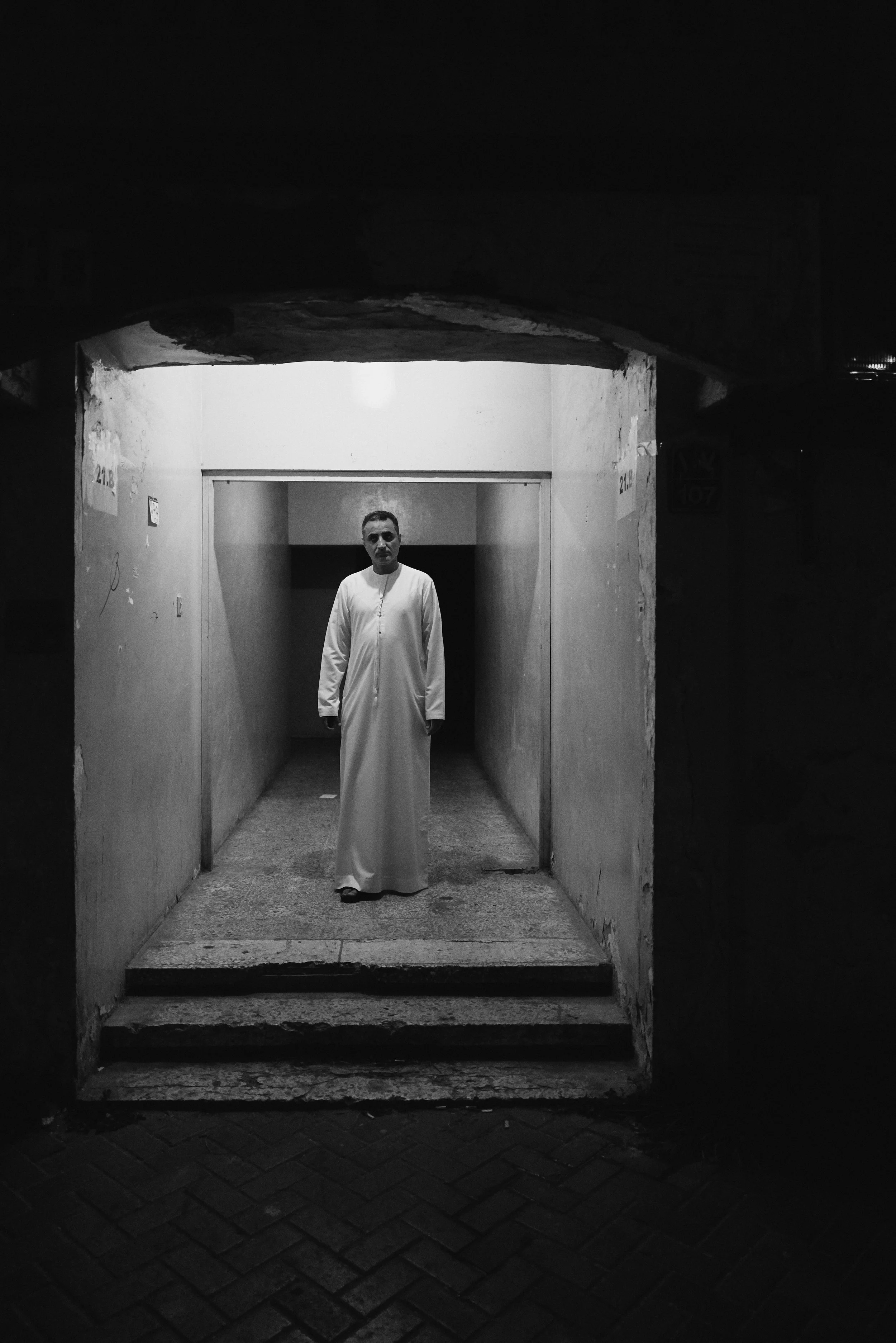 A man dressed in white traditional clothing standing in a dark alleyway entrance with steps leading into a brightly lit interior space.
