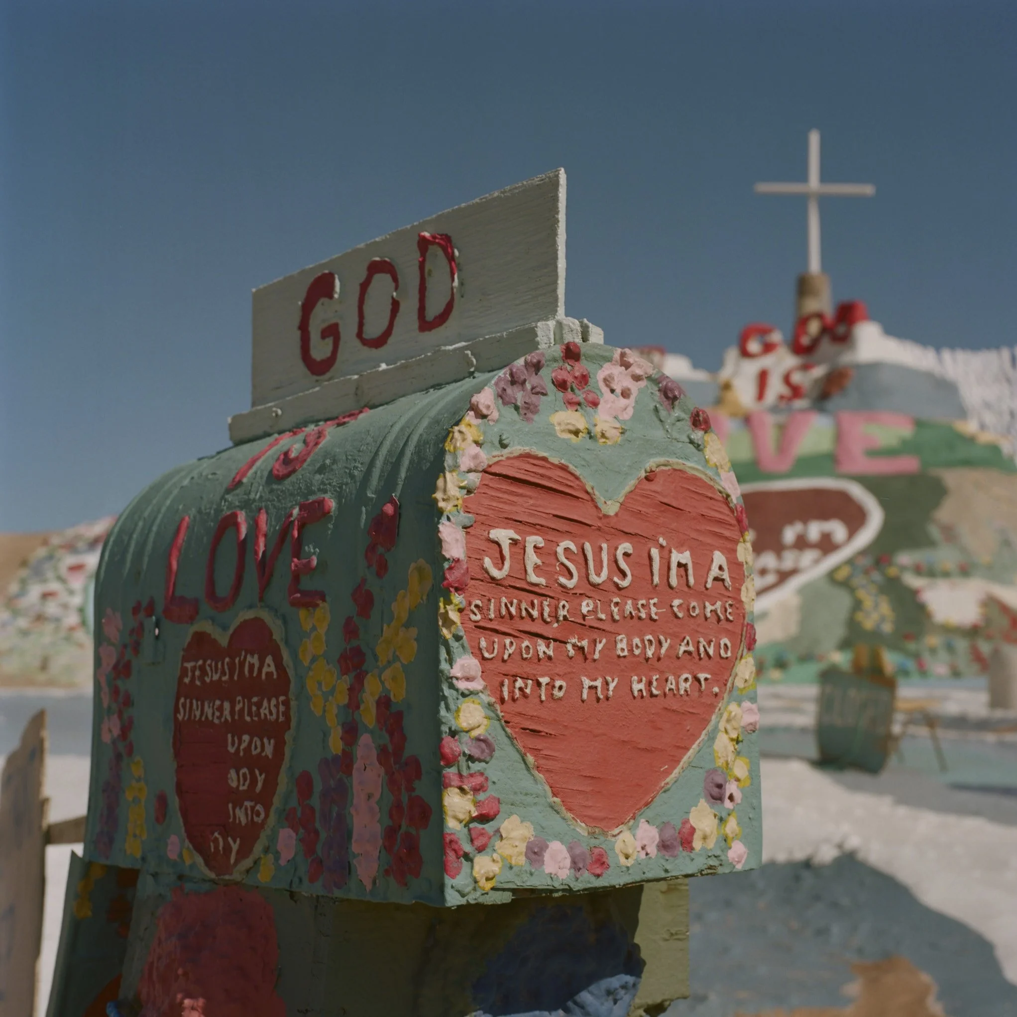 Painted wooden sign with the word 'GOD' in red letters on a white background, surrounded by colorful flowers, with a painted landscape and a church steeple in the background.