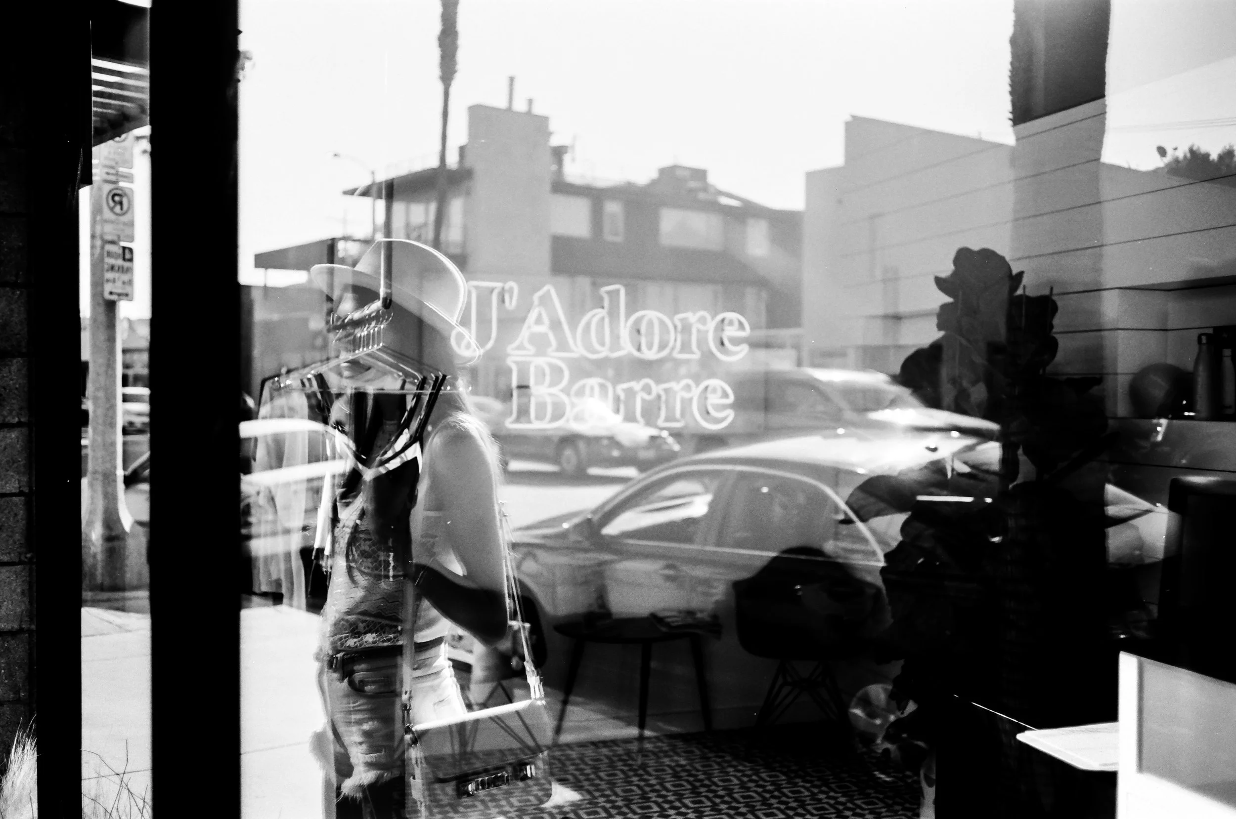 Black and white photo of a person in a hat and patterned clothing taking a selfie in a store window with the store name "Adore Barre" visible reflection including cars and buildings outside.