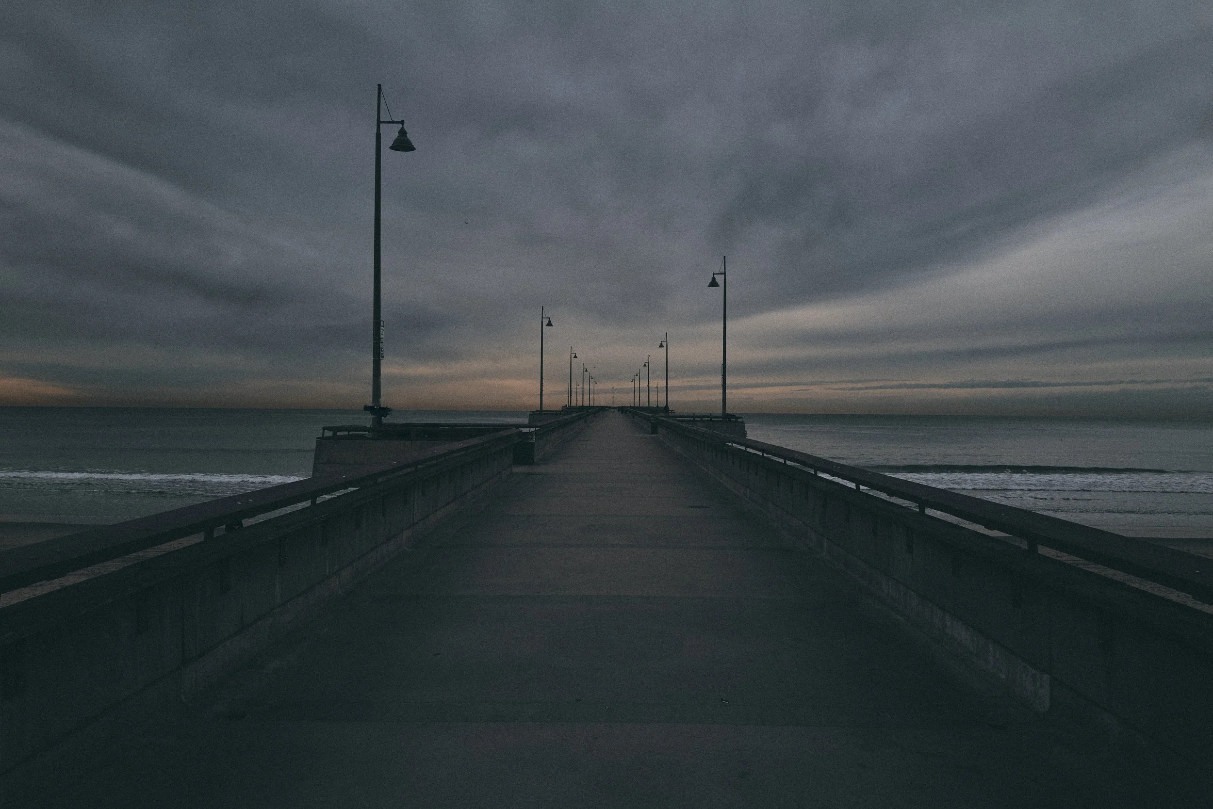 A pier extending into the ocean under a cloudy sky, with tall street lamps along the walkway.