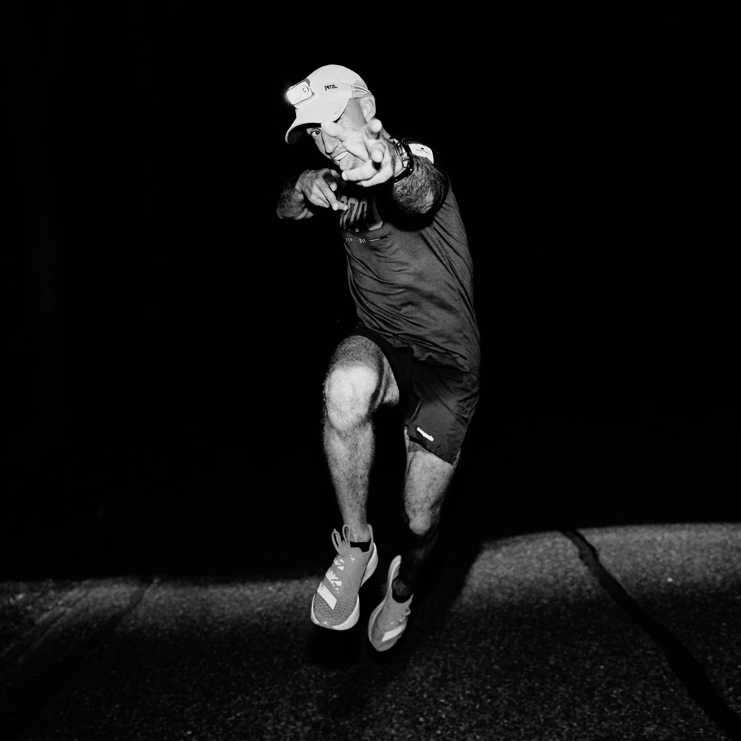 A man wearing a cap with a headlamp, pointing at the camera, in a dark outdoor environment.