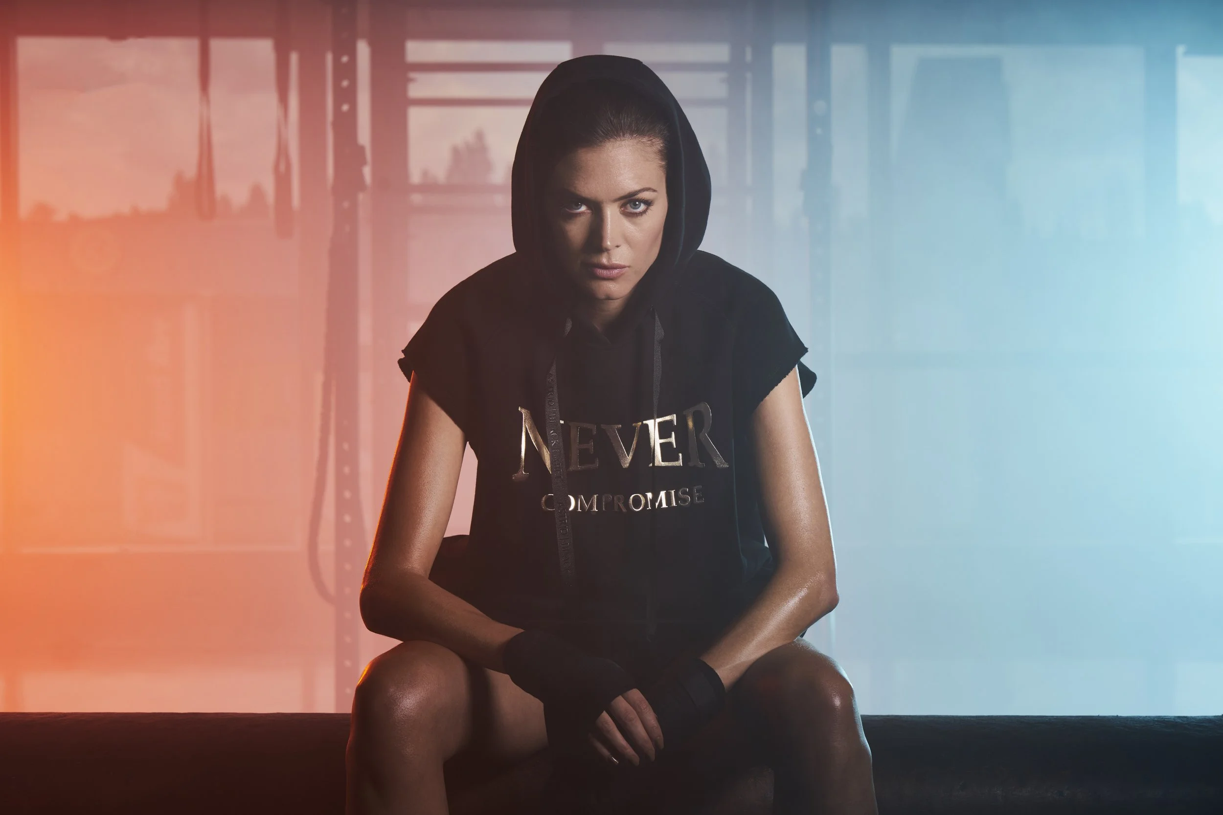 A woman with blue eyes and brown hair tied back, wearing a black hooded shirt with the words 'NEVER PROMISE' on it, sitting with her knees up, in front of a background with orange and blue lighting effects.