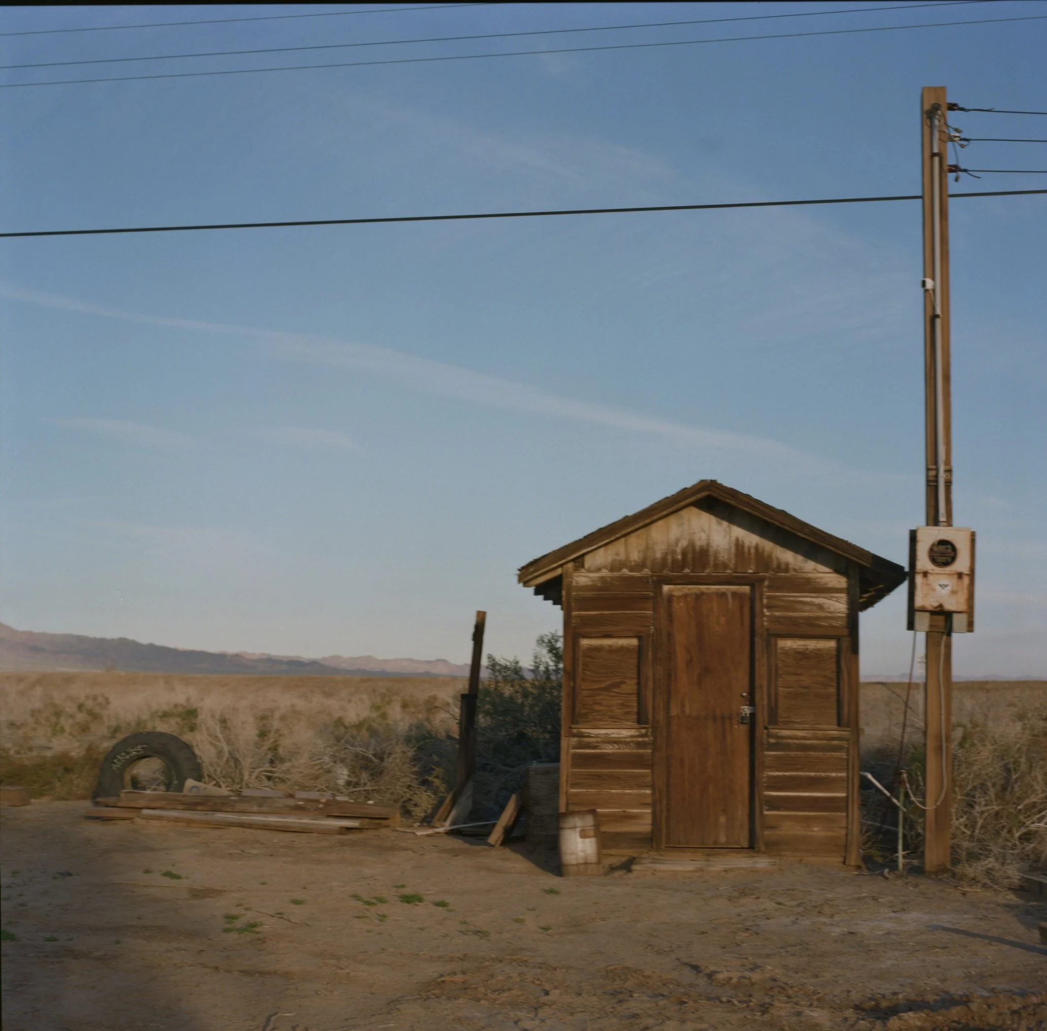 A wooden house with a shingled roof, set in a dry, arid landscape. An electric utility pole with wires extends into the clear blue sky nearby.