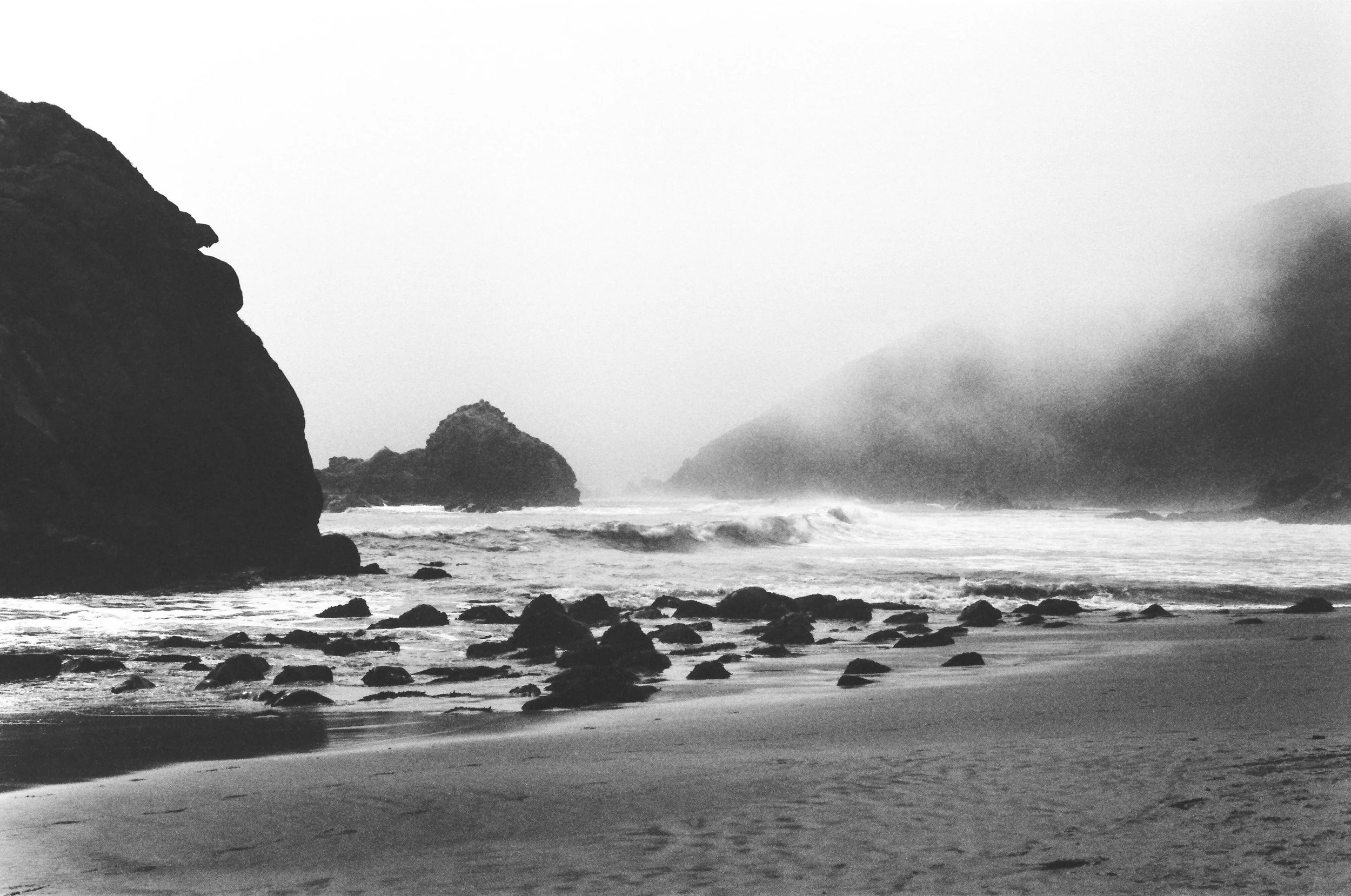 A black-and-white photograph of a rocky beach with waves crashing, large rock formations, and foggy mountains in the background.