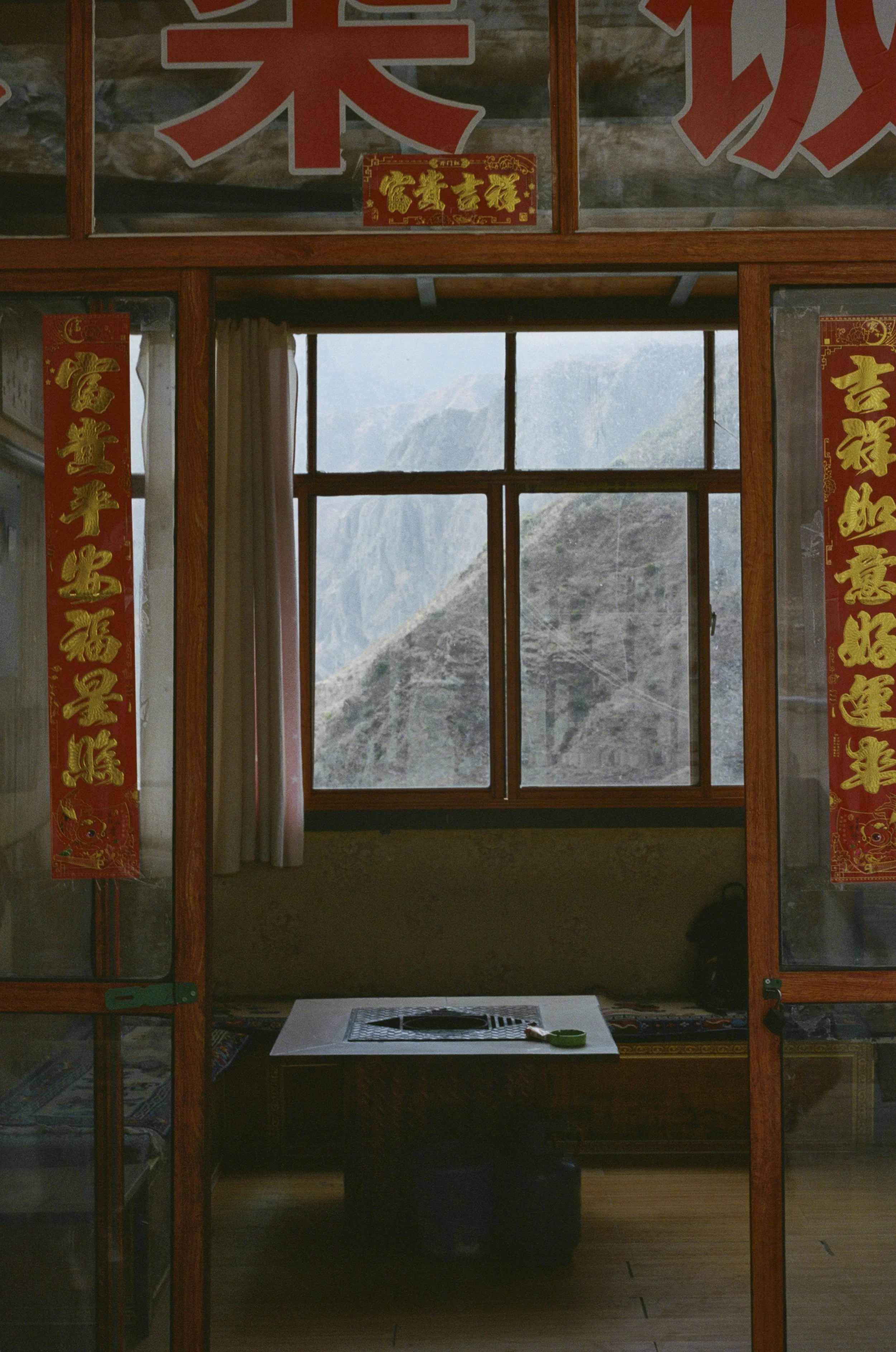 Interior of a room with a window showcasing a mountain view, traditional Chinese couplets hanging on the sides, and a small table with items on it.