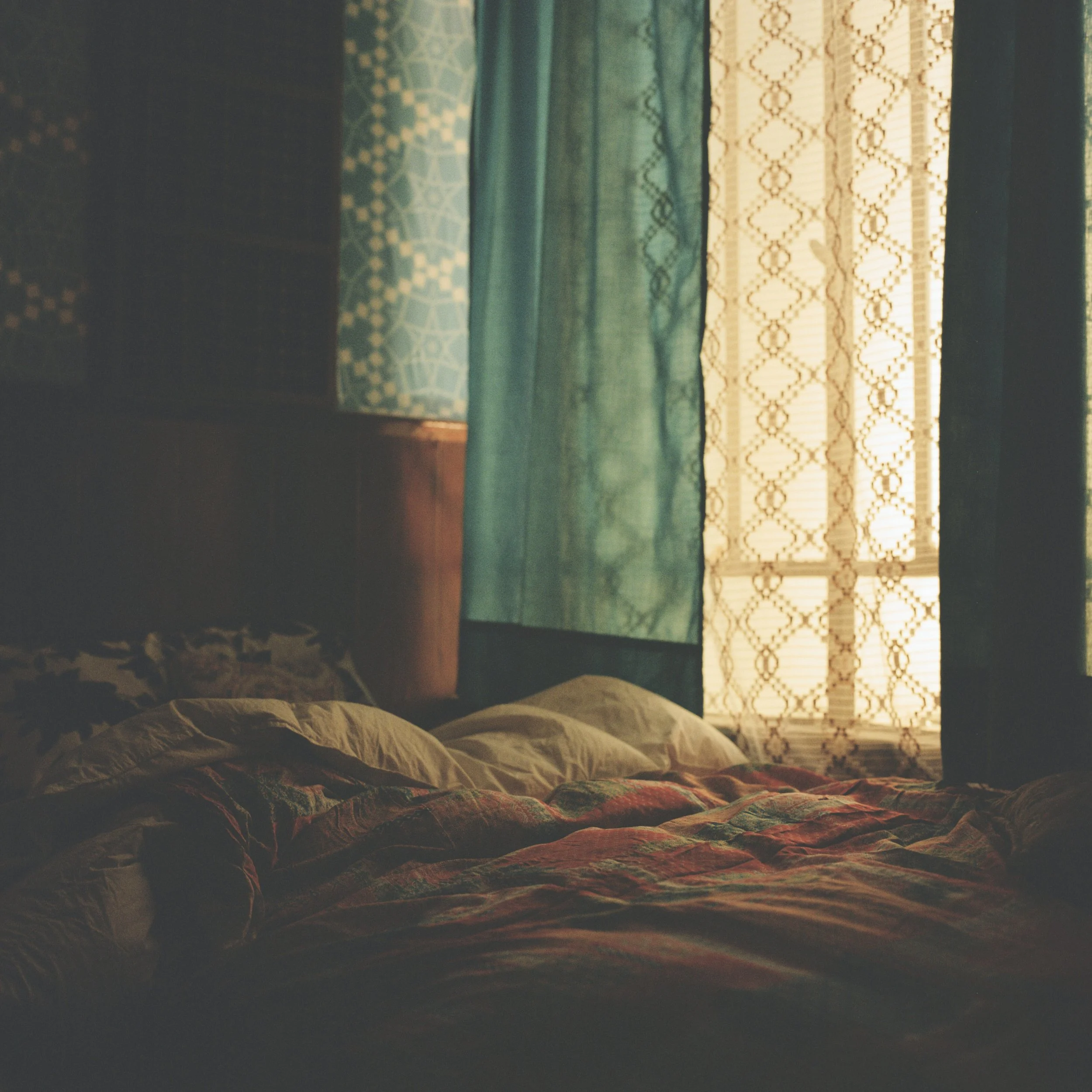 A bed with a patterned quilt and a blanket, with windows in the background covered by curtains with intricate patterns, in a softly lit room.