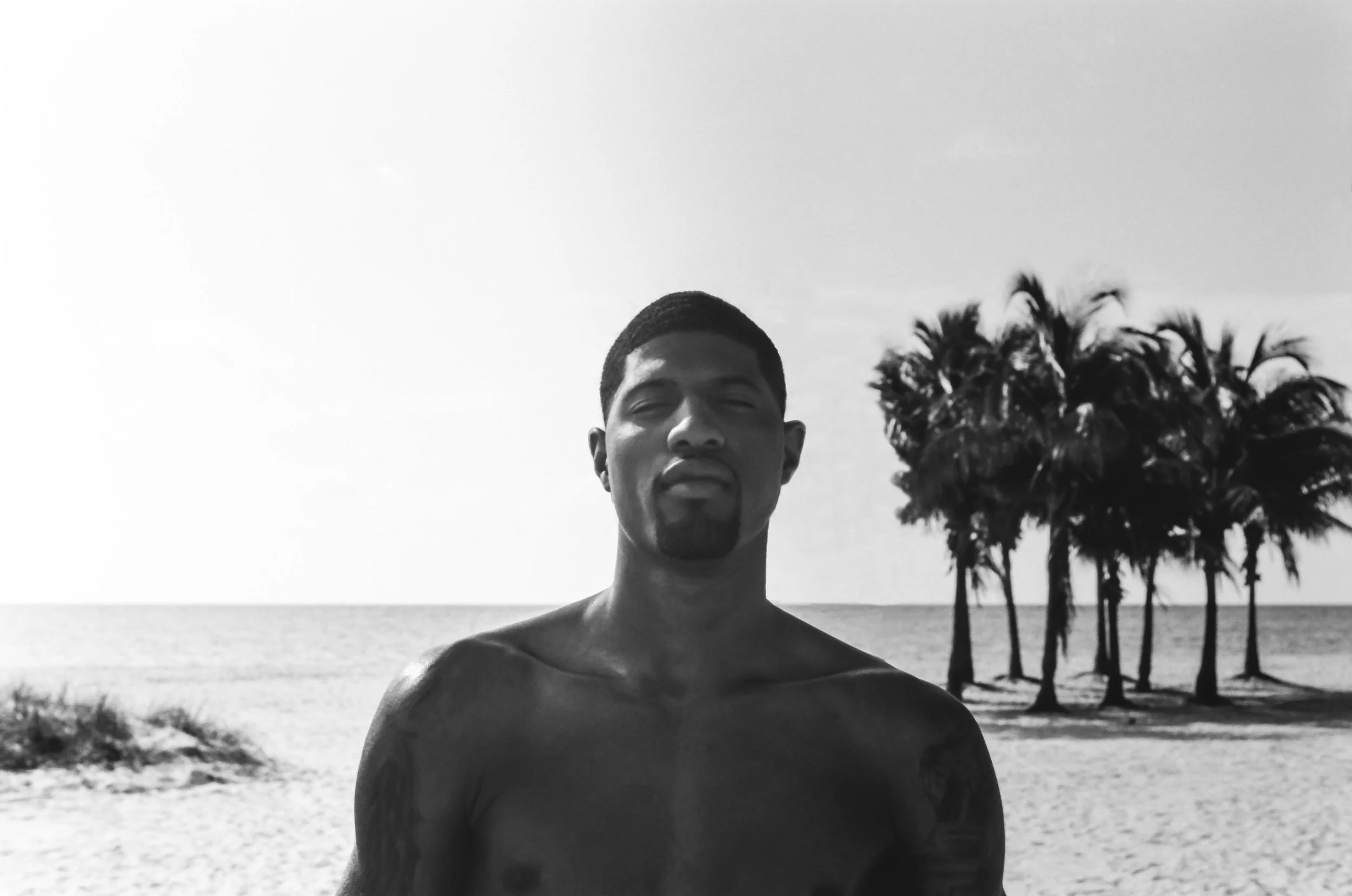 A black and white photo of a shirtless man standing on a sandy beach, with palm trees and the ocean in the background.