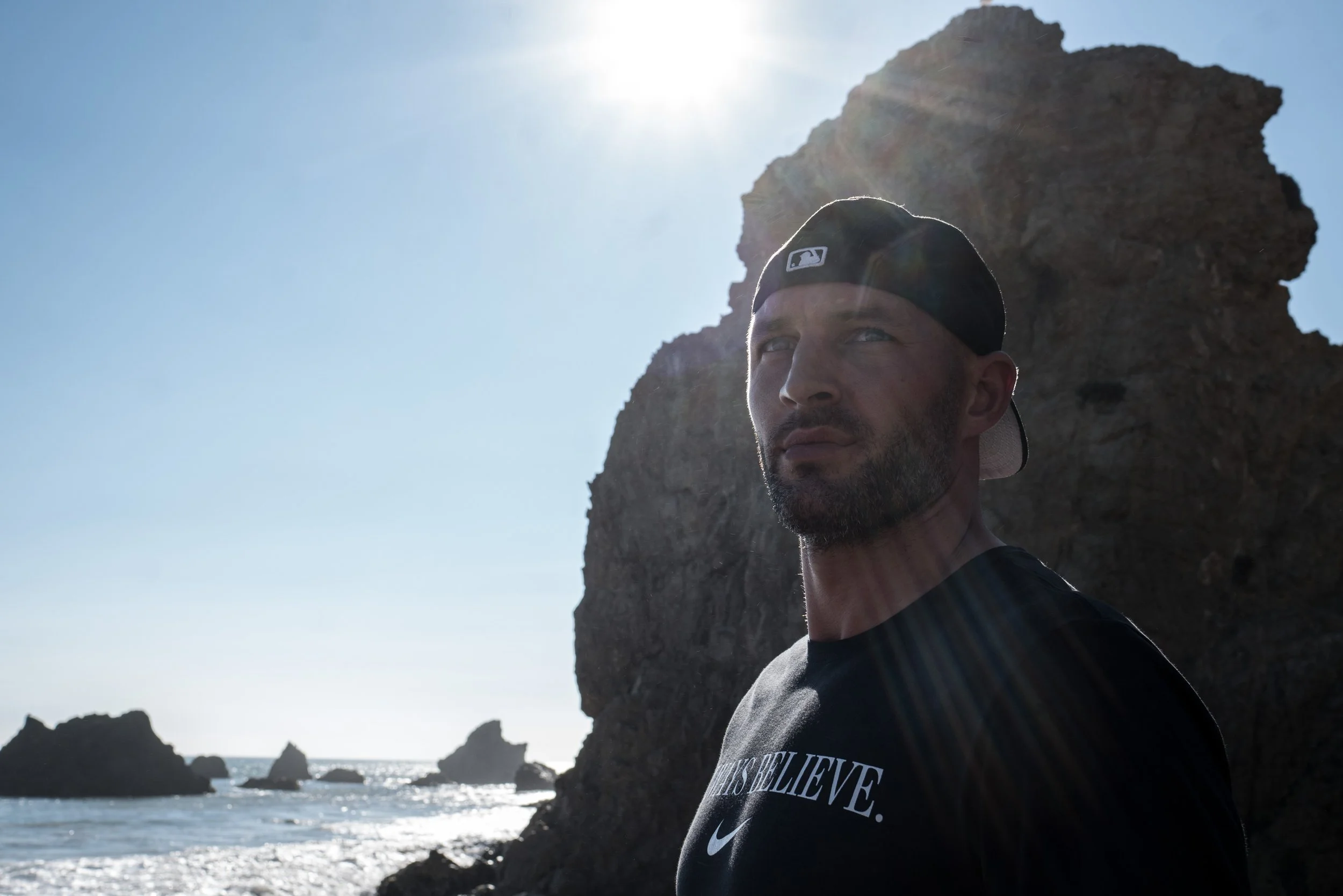 A man with a dark beard and black baseball cap looking into the distance near rocky cliffs by the ocean on a sunny day.