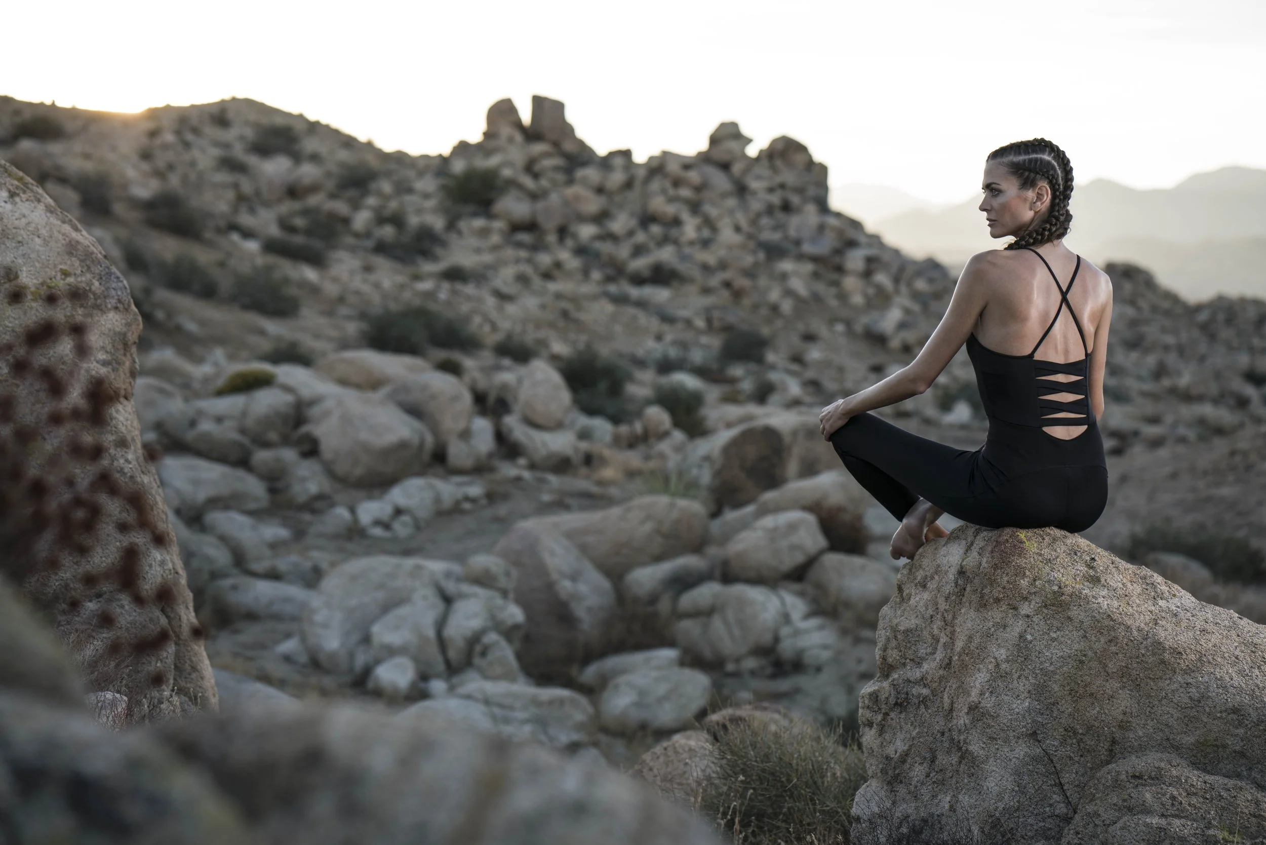 A woman with braided hair practicing yoga on a rock in a rocky desert landscape during sunset.