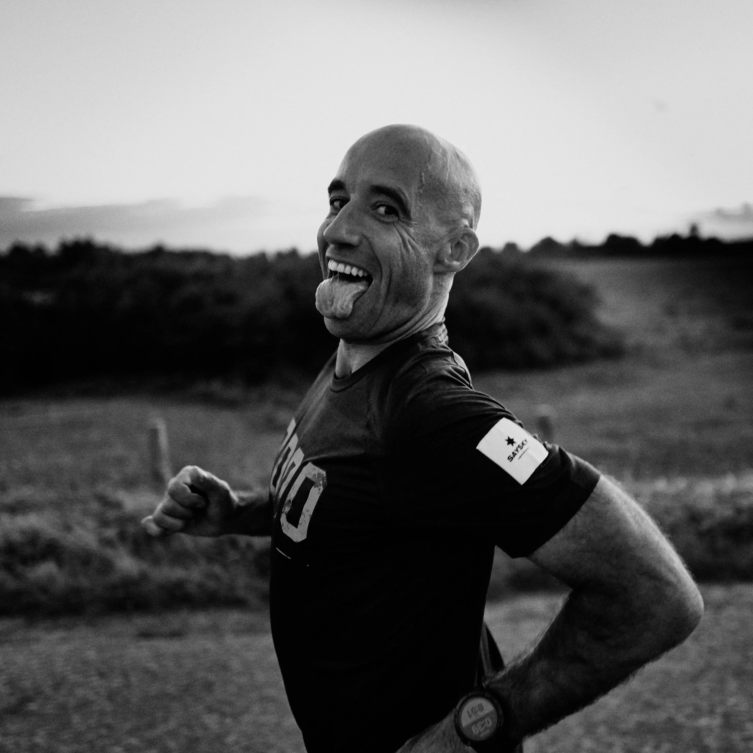 A man with a shaved head and a wide smile sticking out his tongue outdoors in a field, wearing a sports jersey with the number 23.