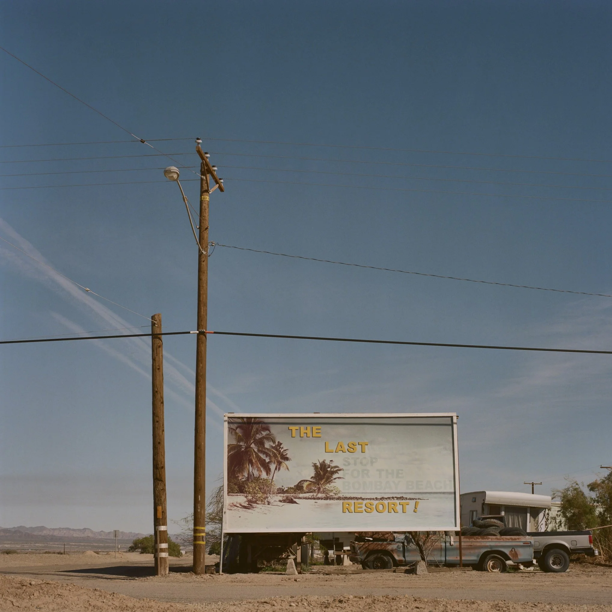 A roadside sign advertising a resort near Bombay Beach with palm trees and a desert landscape in the background.