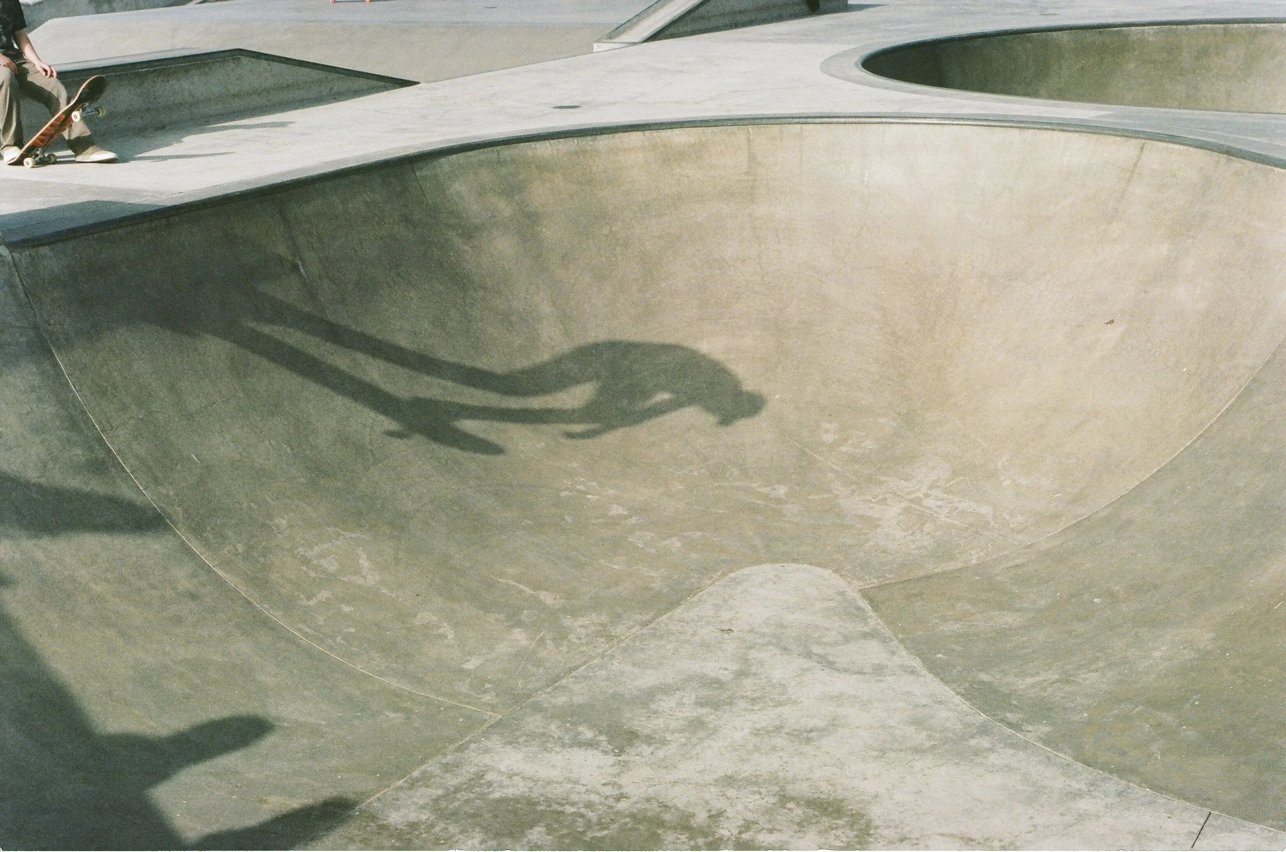 A skate park with empty concrete bowls and ramps. There is a person sitting on the edge of a bowl with a skateboard leaning next to them. The shadows cast by the skateboards and the person are visible on the concrete.