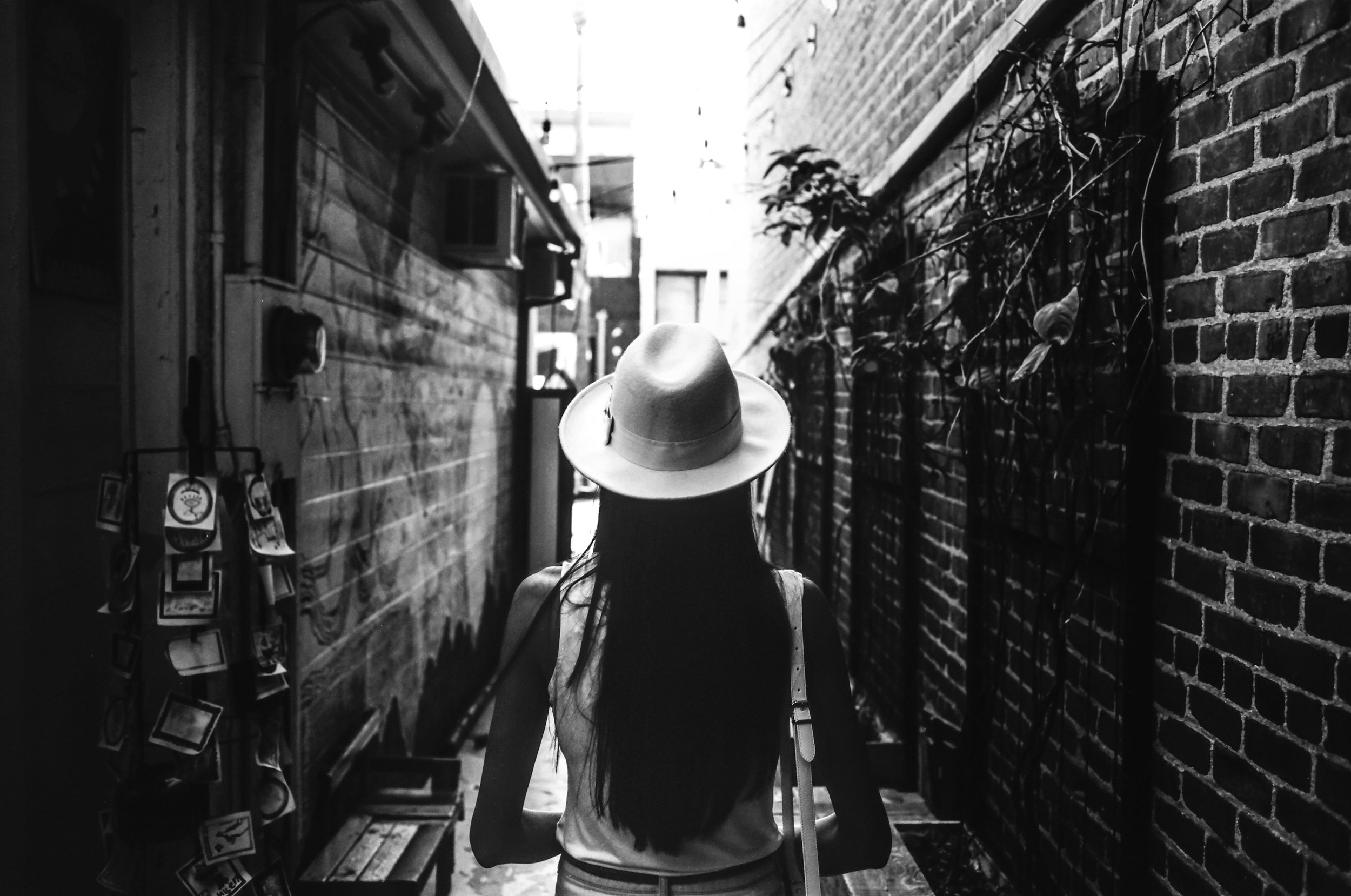 A person wearing a hat and a sleeveless top walking through a narrow alley with brick walls on either side, one with vines growing on it, in black and white.