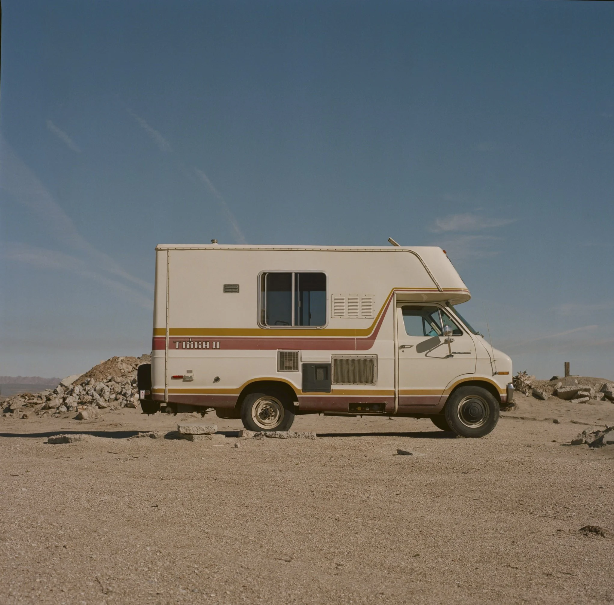 A white motorhome with orange and yellow stripes parked on a dirt area with rocks, under a clear blue sky.