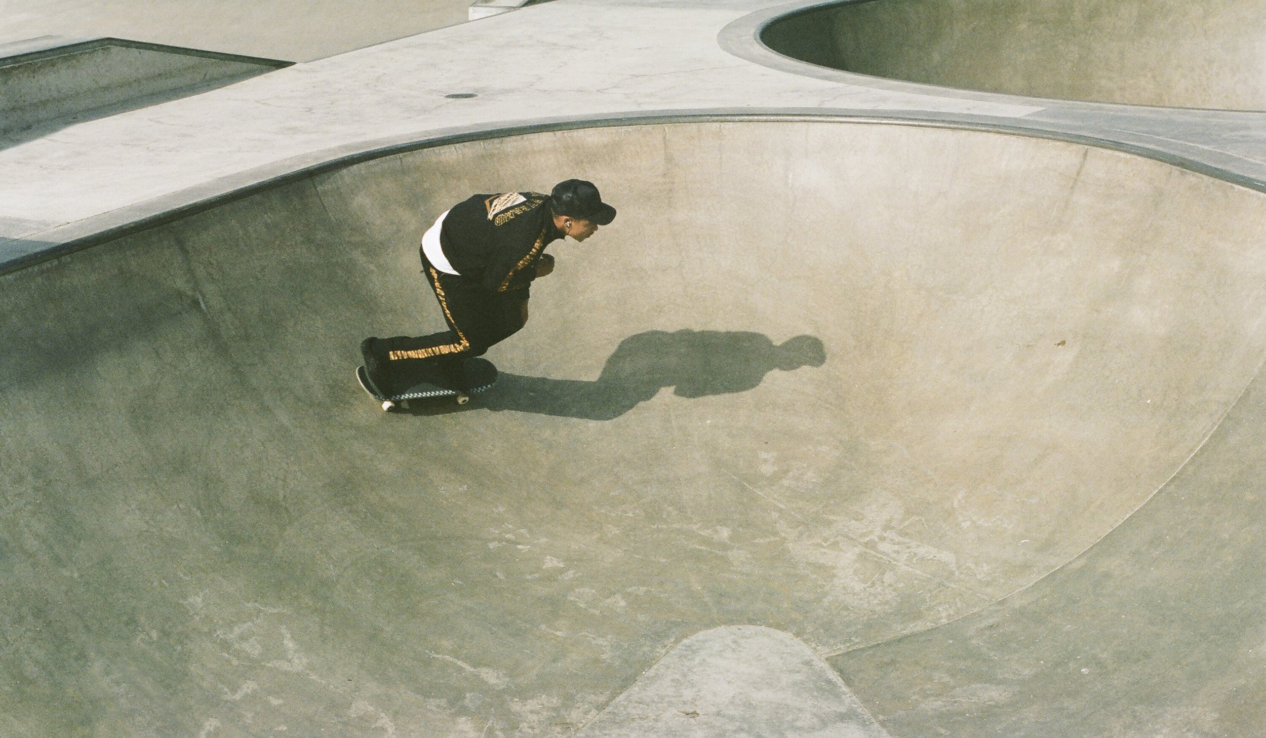 A woman in black with yellow patterns on her sleeves is bending over on a skateboard in a concrete skate park, with her shadow cast on the bowl.