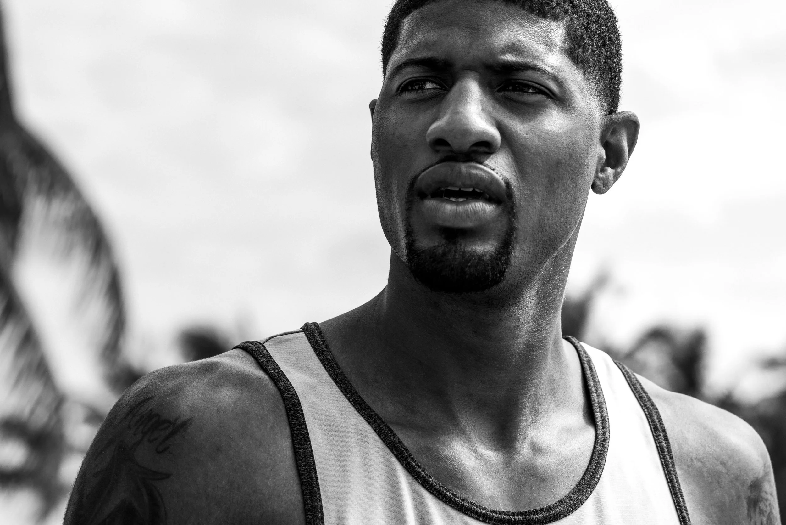 A black and white close-up of a young man with short hair, a goatee, and a tattoo on his arm, wearing a sports jersey, looking to the side with a slightly open mouth.