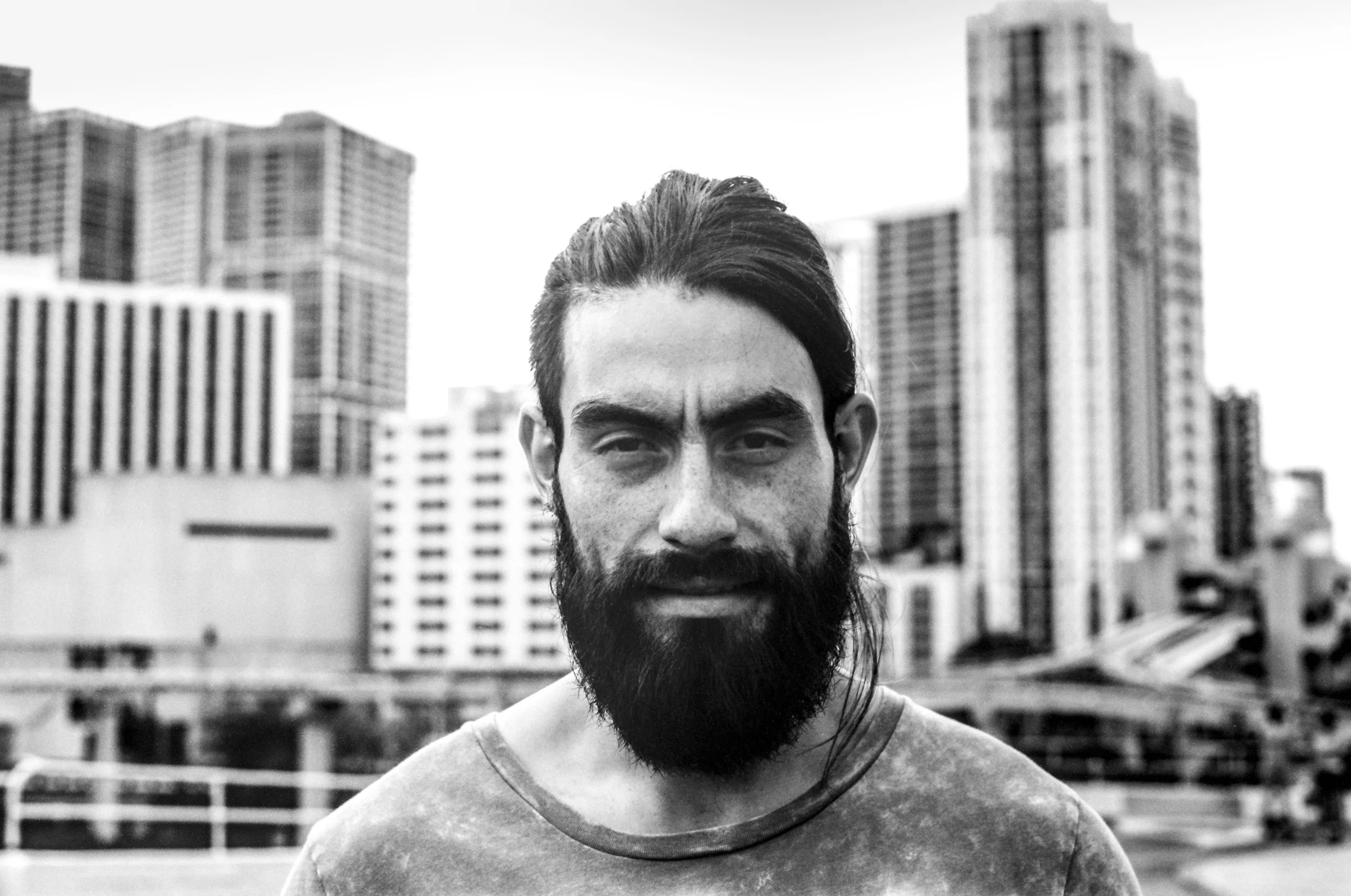 A man with a beard and short hair smiling in front of a city skyline with tall buildings, in black and white.