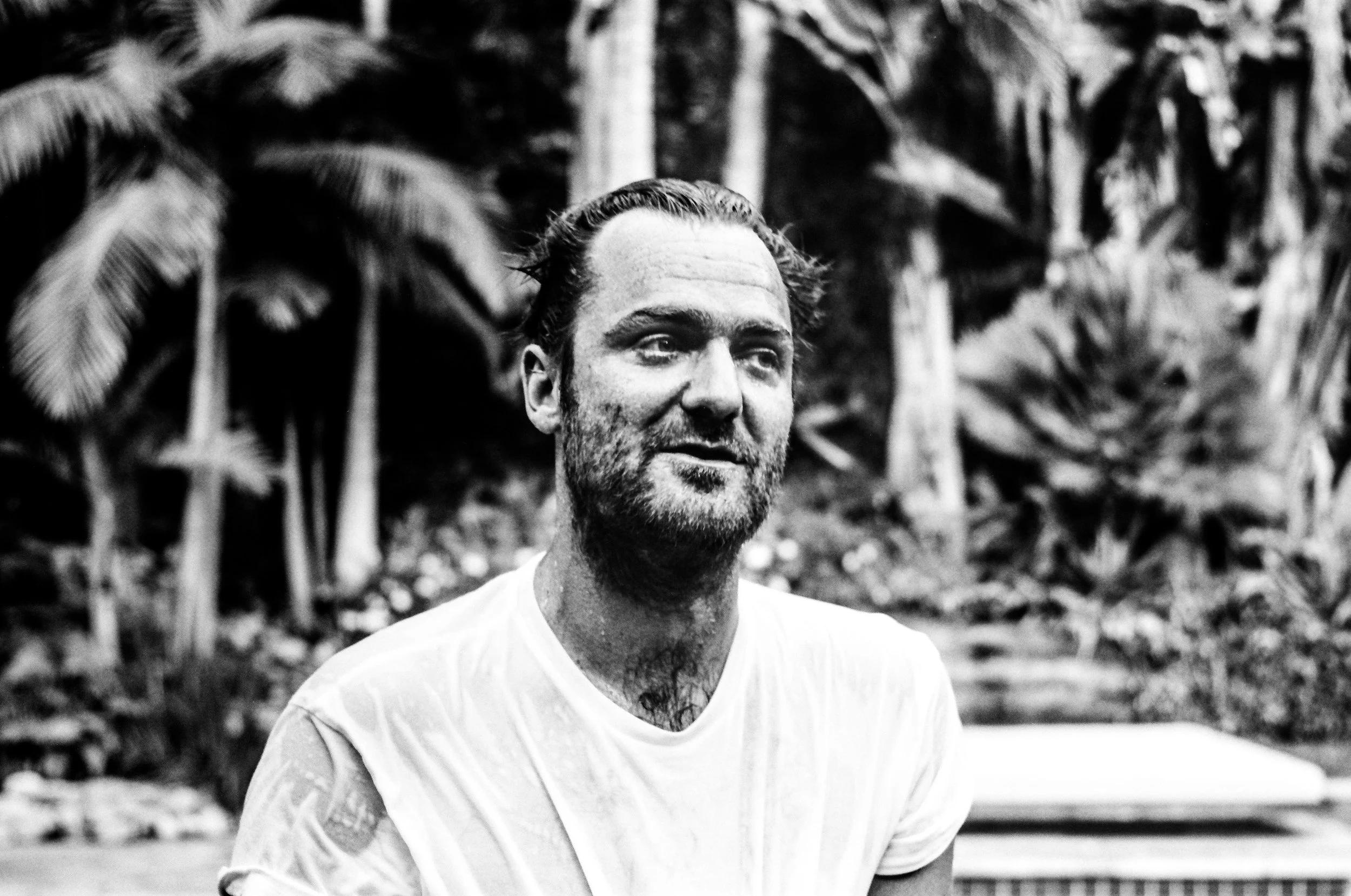 A black-and-white photograph of a man with a beard and mustache, smiling slightly, outdoors with trees in the background.