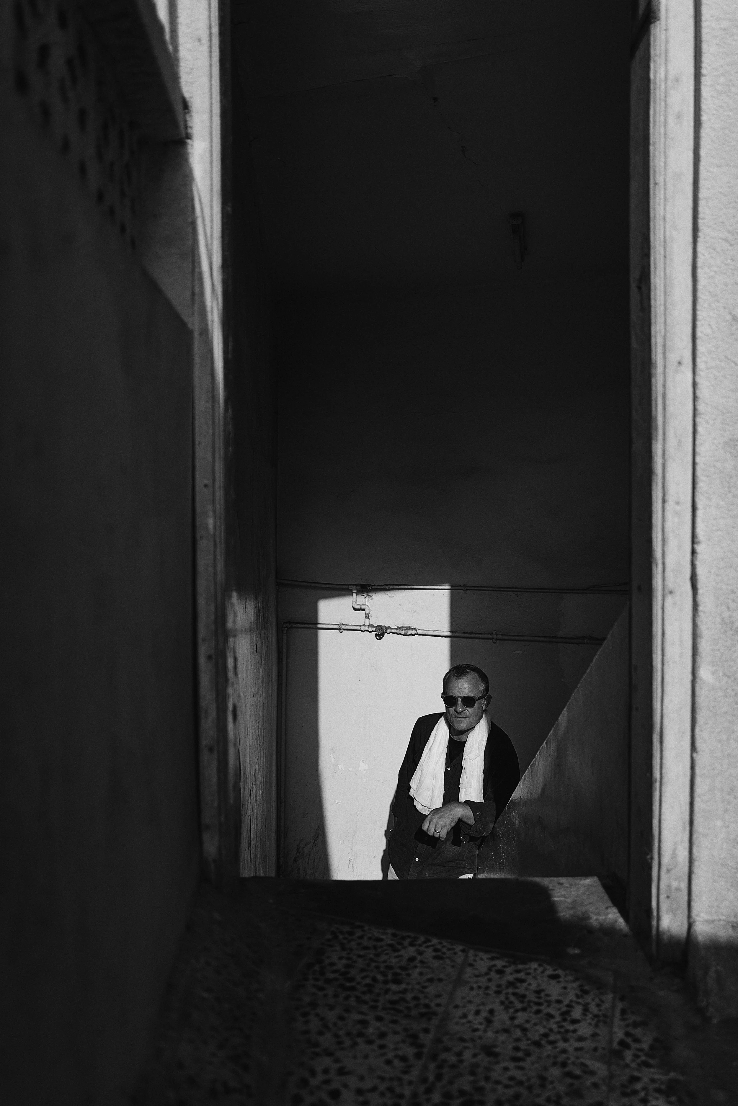 Black and white photo of a man wearing sunglasses and a white scarf, standing in an outdoor corridor or alley with shadows cast on the wall.